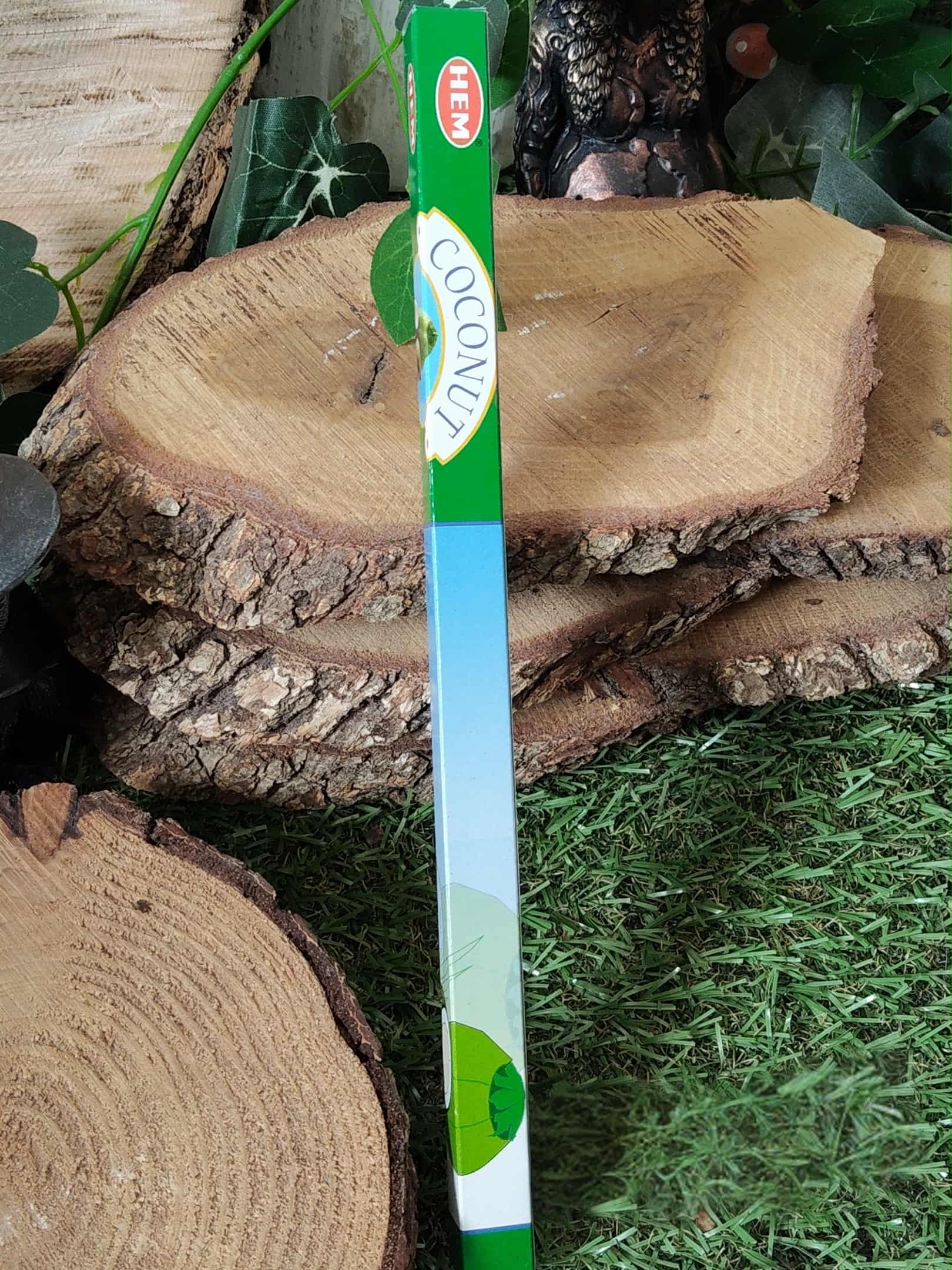 Boîte d’encens noix de coco HEM verticale entre rondins de bois et gazon — parfum tropical doux pour une ambiance relaxante et chaleureuse.