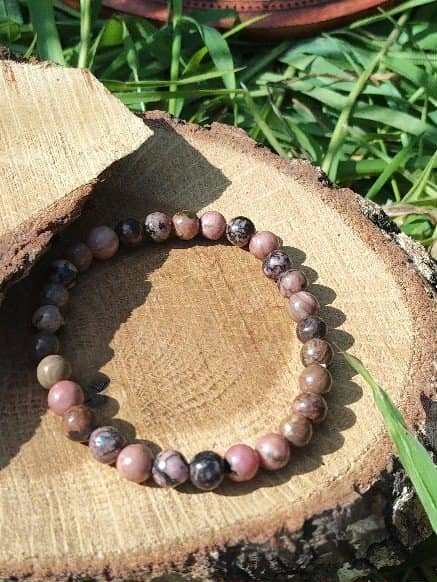 Bracelet rhodonite 6 mm sur rondin de bois au soleil avec herbes vertes, pierre naturelle pour réconfort émotionnel, tendresse intérieure et apaisement du cœur