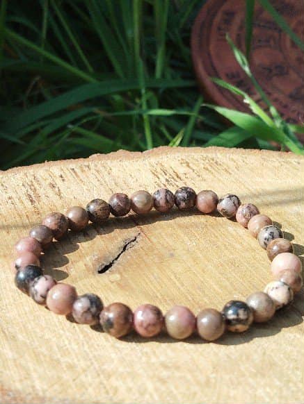 Bracelet rhodonite 6 mm sur support en bois avec herbes en fond et lumière chaude, confiance retrouvée, stabilité du cœur et douceur relationnelle