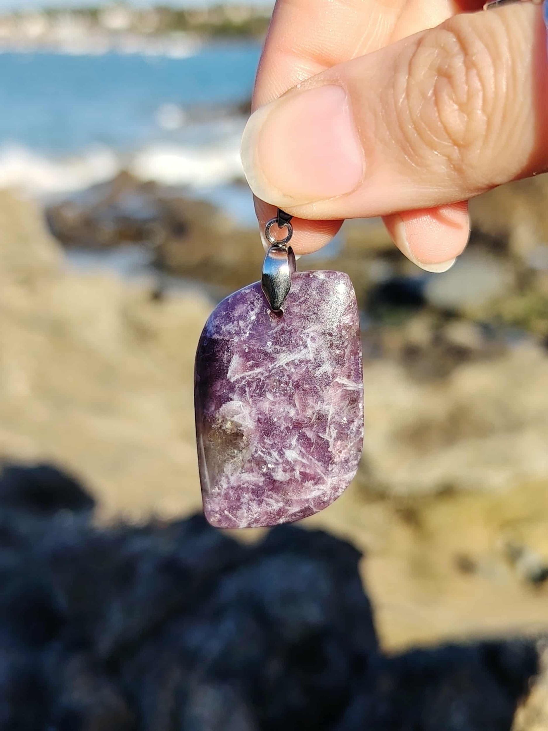 PPendentif lépidolite rectangulaire tenu à la main en bord de mer, bijou en pierre naturelle évoquant le lâcher-prise, la tendresse et la paix du mental.