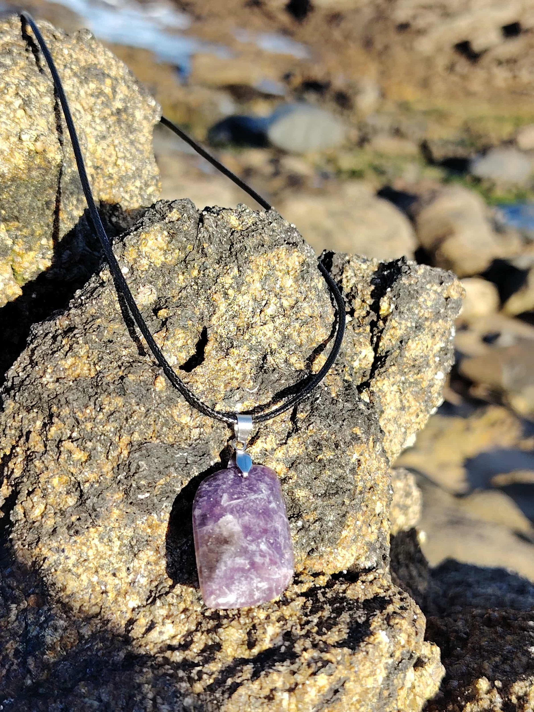 Pendentif lépidolite rectangulaire accroché sur rocher en bord de mer en Bretagne, bijou en pierre naturelle lié à la sérénité, au relâchement intérieur et à la souplesse émotionnelle.