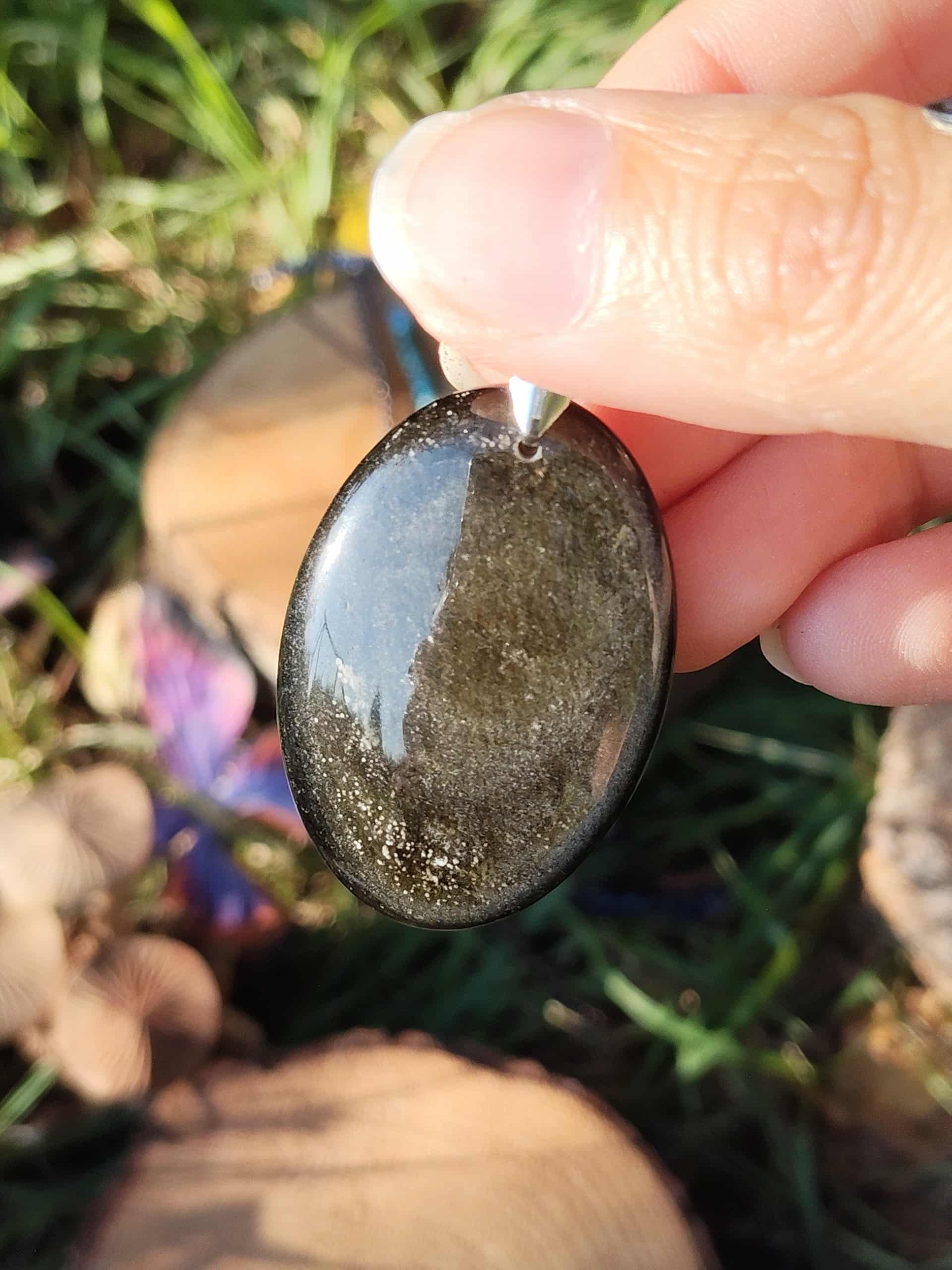 Pendentif ovale en obsidienne dorée avec éclats dorés, présenté dans la main en décor nature, pierre naturelle reliée au discernement, à la confiance calme et à la présence à soi.