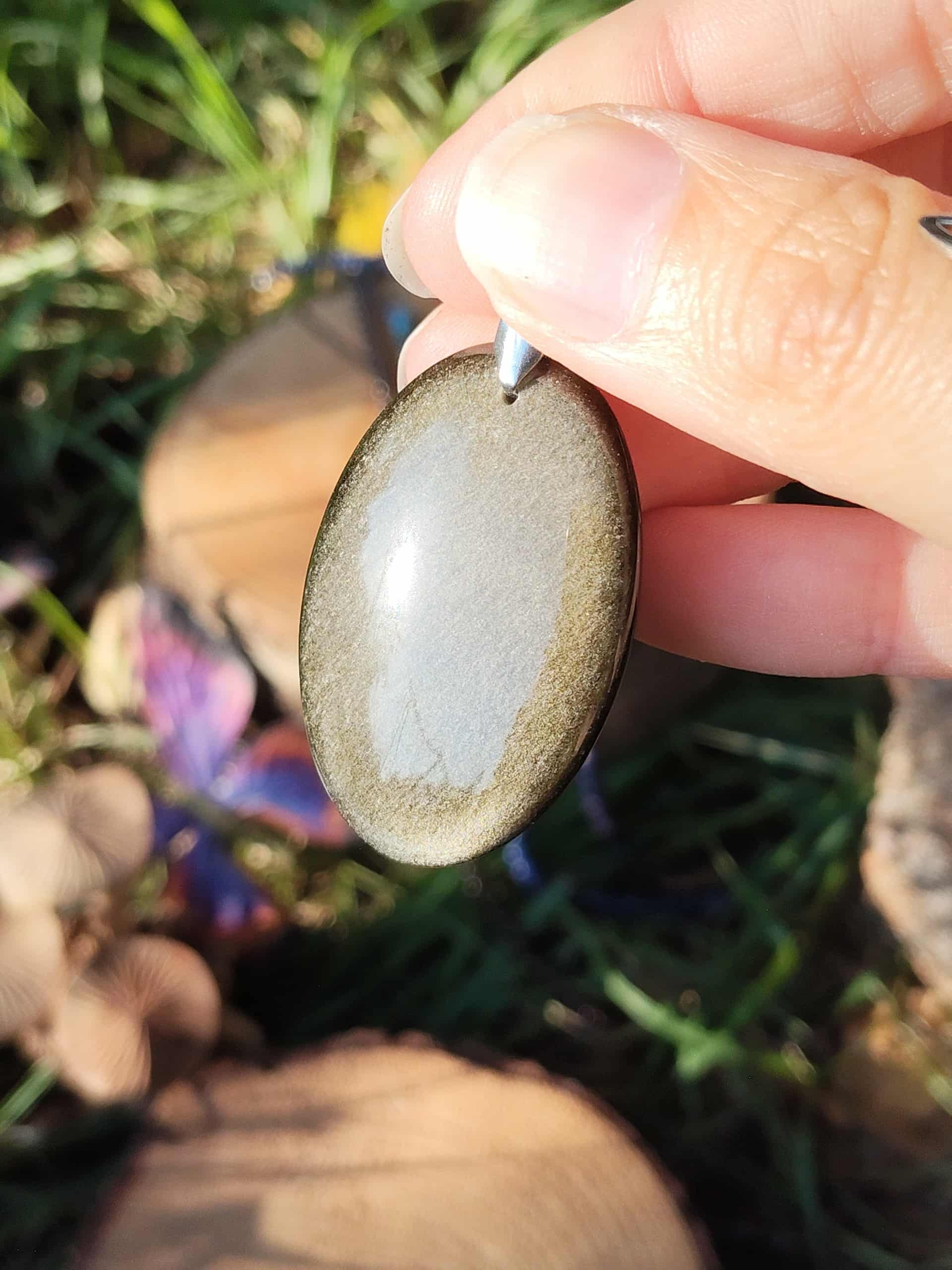 Pendentif obsidienne dorée tenu dans la main sur fond d’herbe et de fleurs, bijou en pierre naturelle associé à l’enracinement, à la solidité intérieure et à la sérénité.