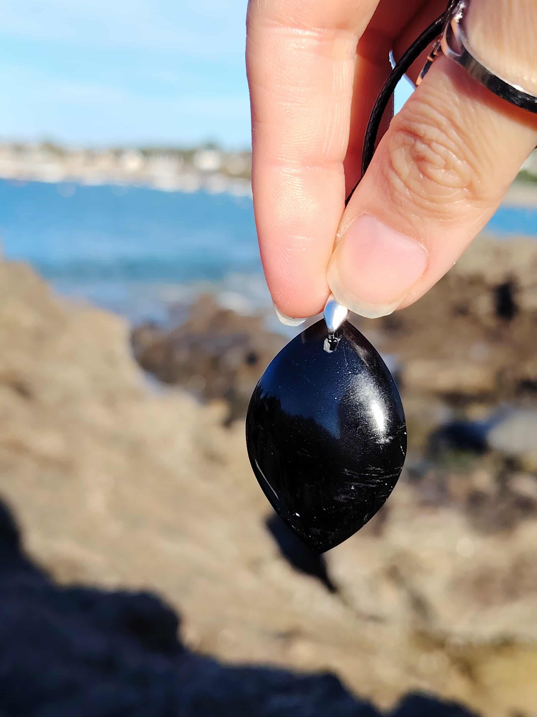 Pendentif tourmaline noire marquise tenu entre les doigts face à la mer en Bretagne, bijou en pierre naturelle évoquant l’ancrage, la protection et la maîtrise de soi.