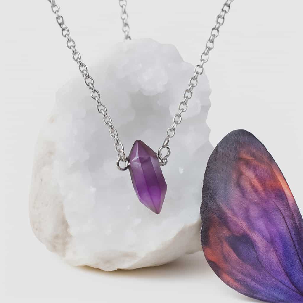 Pendentif pointe améthyste naturelle sur chaîne acier inoxydable, présenté sur amas de quartz et aile violette, collier pierre violette lié à l’apaisement, à l’harmonie émotionnelle et à l’élévation spirituelle.