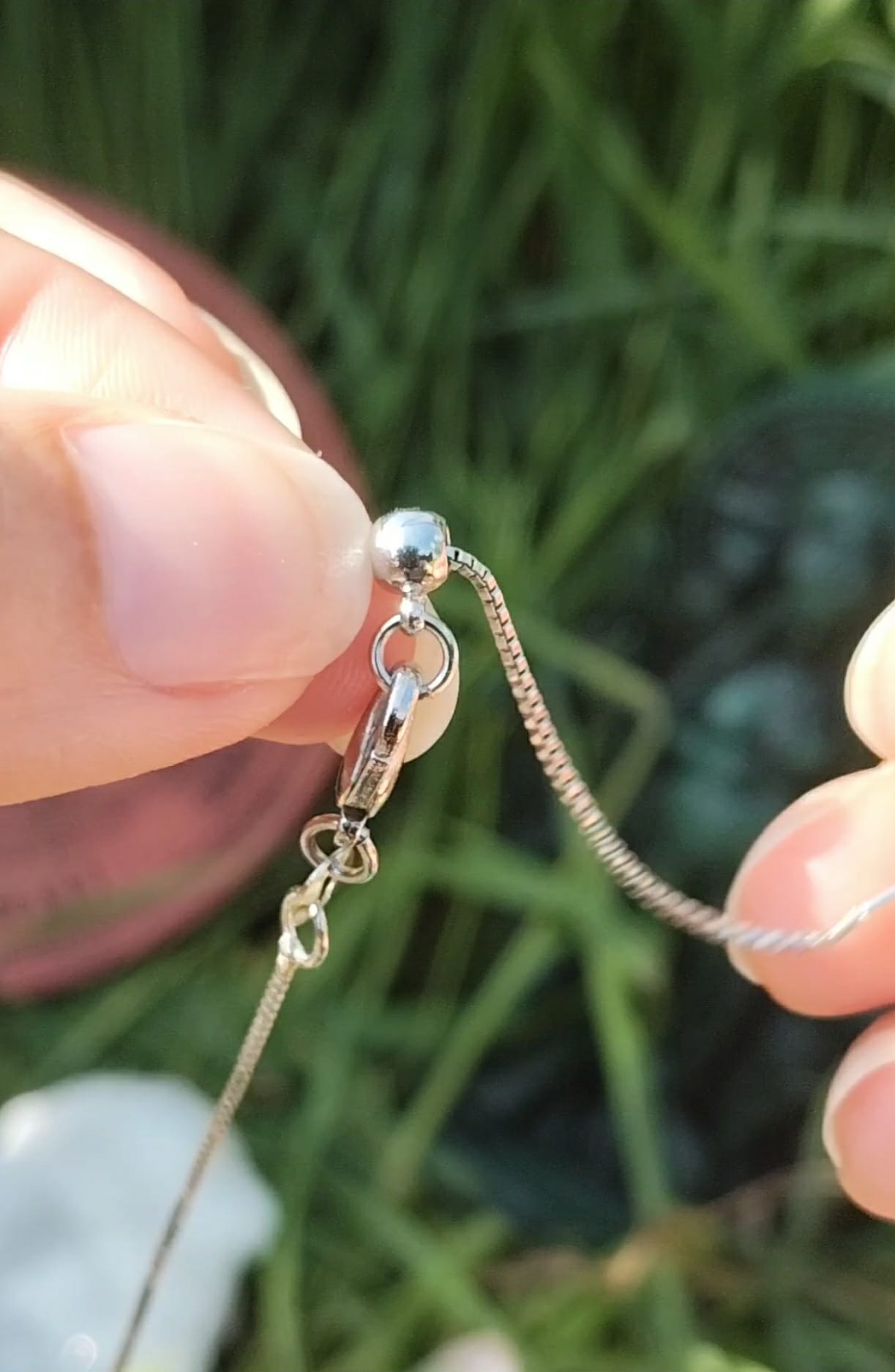Gros plan sur chaînette réglable du pendentif goutte améthyste tenue entre les doigts, plaqué argent sur fond d’herbe