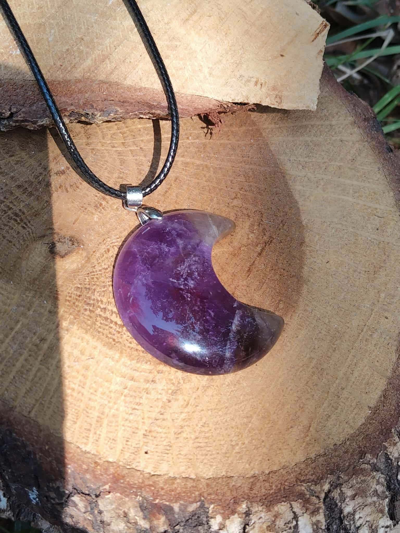Pendentif lune améthyste sur tranche de bois en extérieur, cordon noir et décor végétal, bijou en pierre naturelle lié à l’apaisement, au lâcher-prise et à l’écoute de soi.