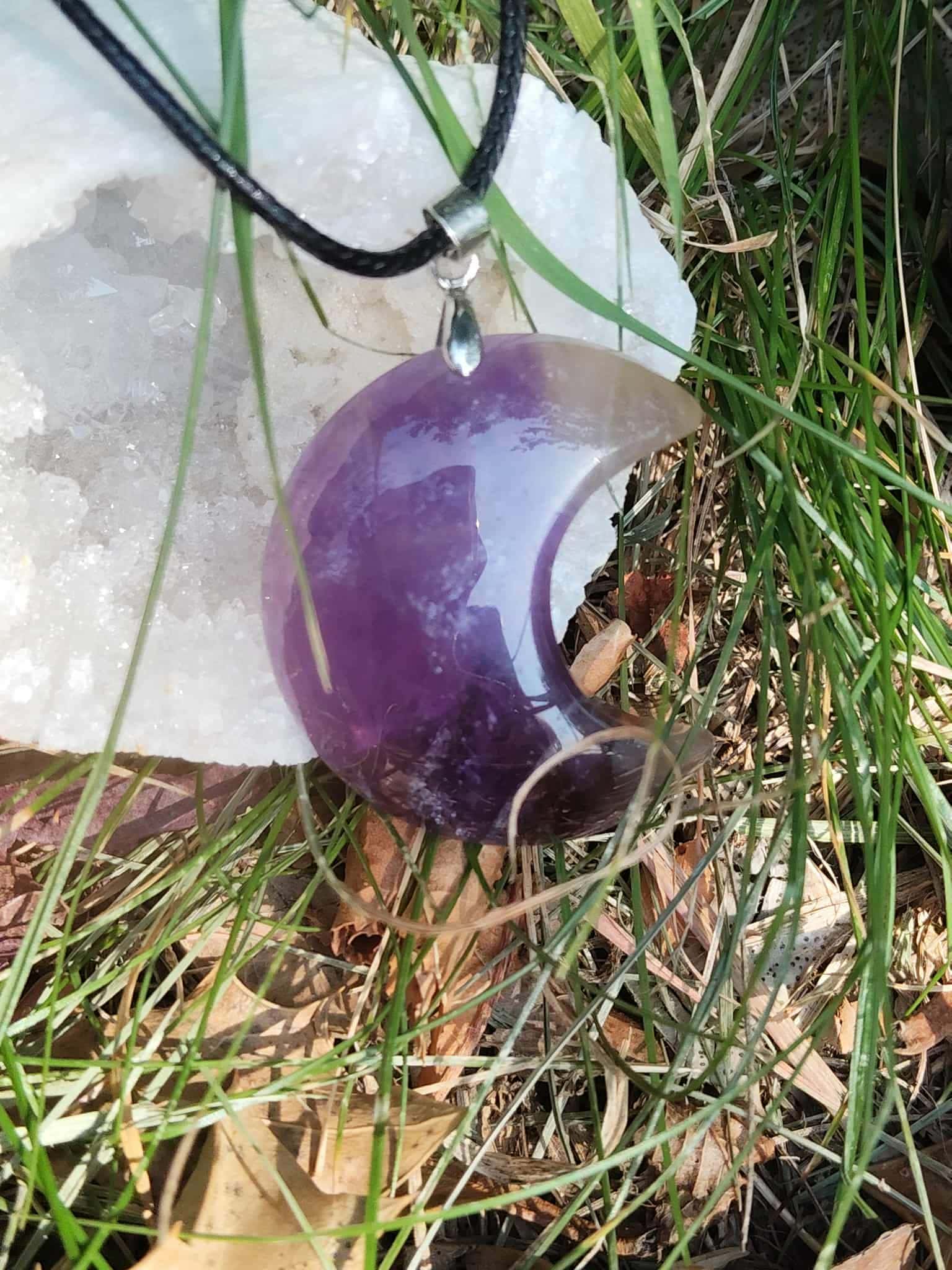 Pendentif lune en améthyste dans l’herbe contre un amas de quartz, cordon noir visible, bijou naturel associé à la paix intérieure, à l’intuition et au réconfort émotionnel.
