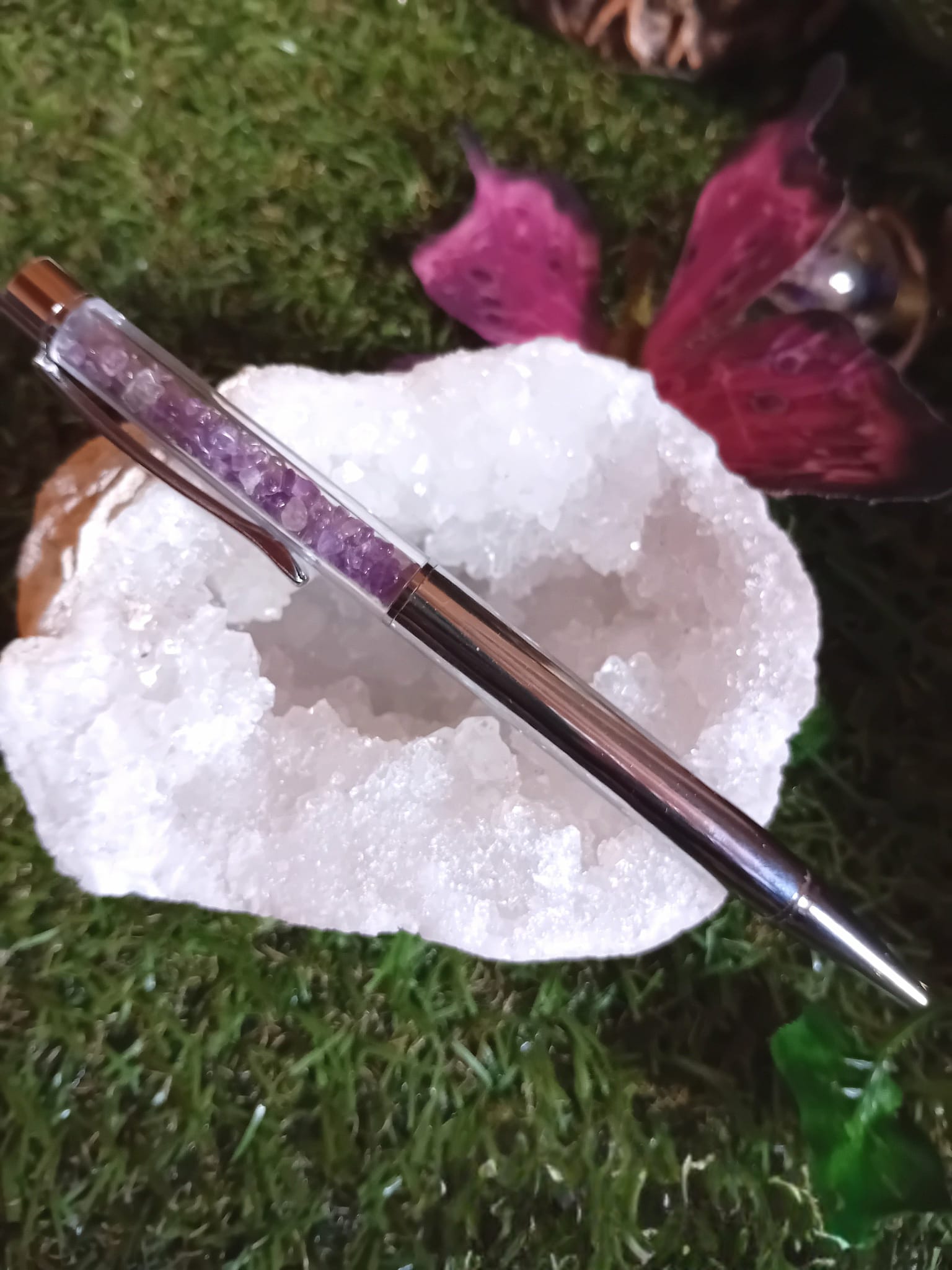 Stylo bille rempli d’éclats d’améthyste posé sur une grande druse de cristal blanc avec pétales de rose sur l’herbe, accessoire d’écriture en pierres naturelles pour créativité et apaisement du mental.