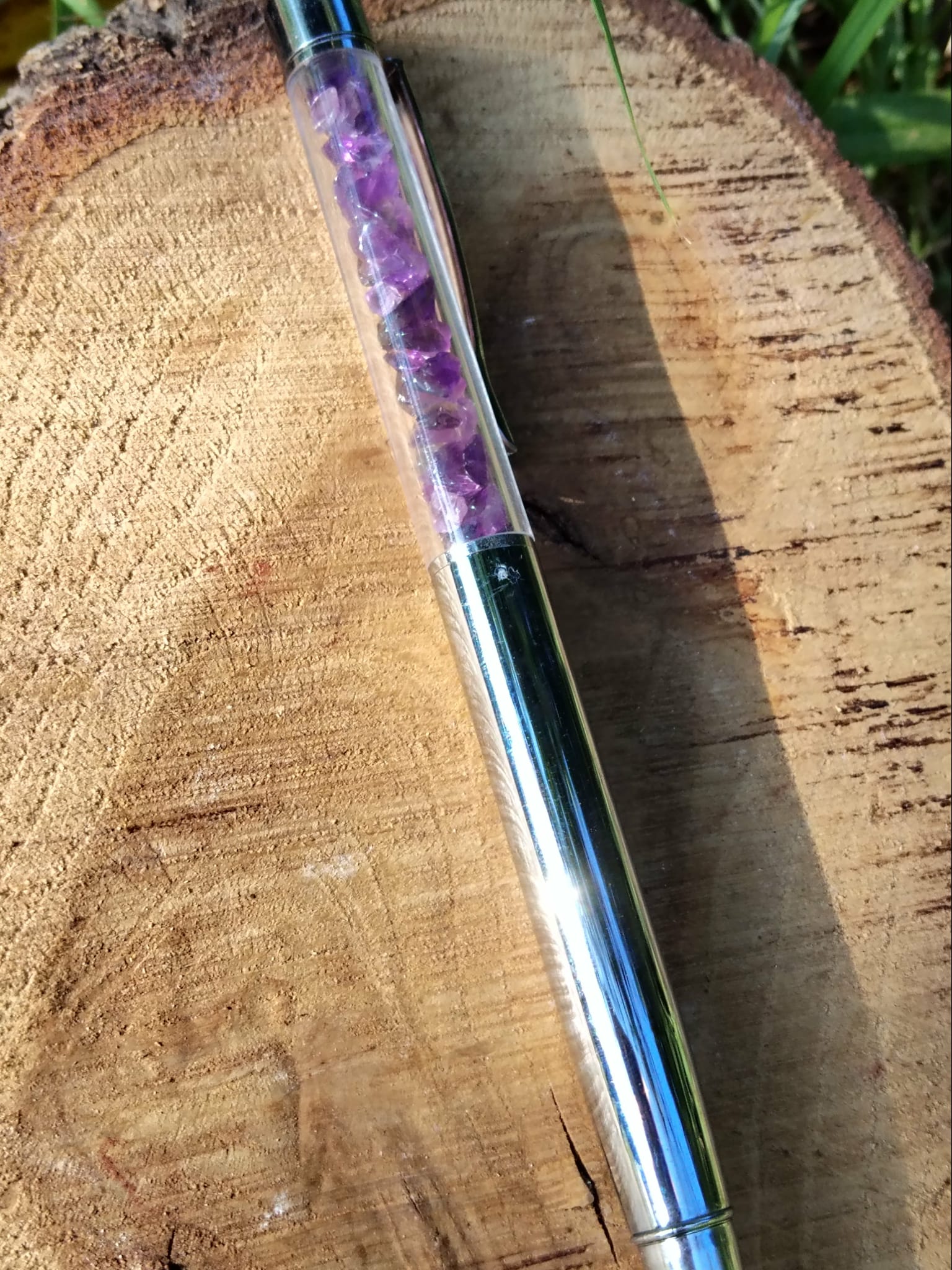 Stylo améthyste rechargeable posé sur une tranche de bois en extérieur, tube transparent rempli d’éclats violets, écriture fluide et design élégant.