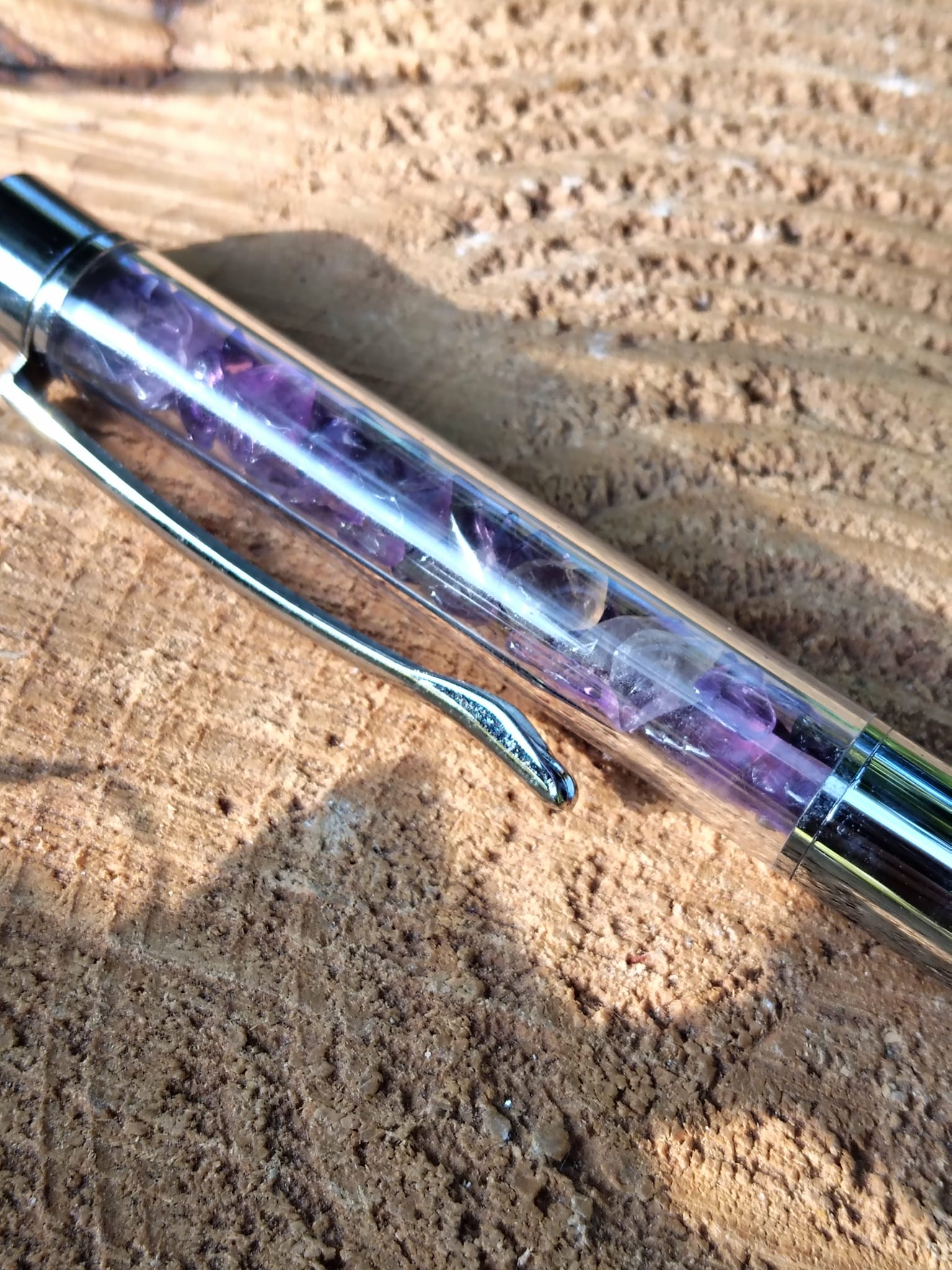 Stylo bille transparent rempli d’éclats d’améthyste violette en gros plan, posé sur une surface de bois, accessoire d’écriture en pierres naturelles pour créativité, inspiration et apaisement du mental.