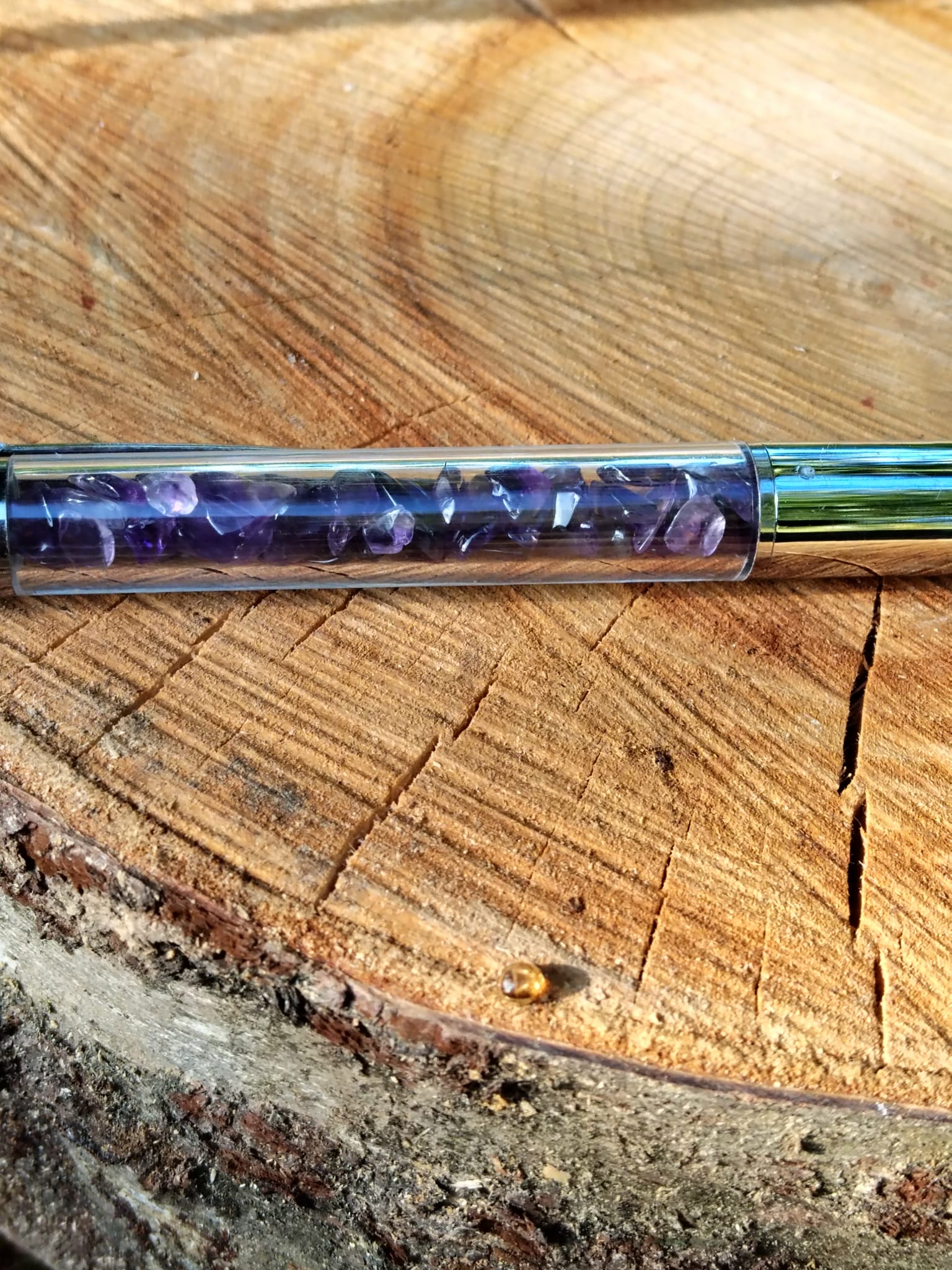 Gros plan sur le réservoir d’un stylo bille rempli d’éclats d’améthyste violette, posé sur une rondelle de bois, accessoire d’écriture en pierres naturelles pour inspiration, concentration et apaisement du mental.