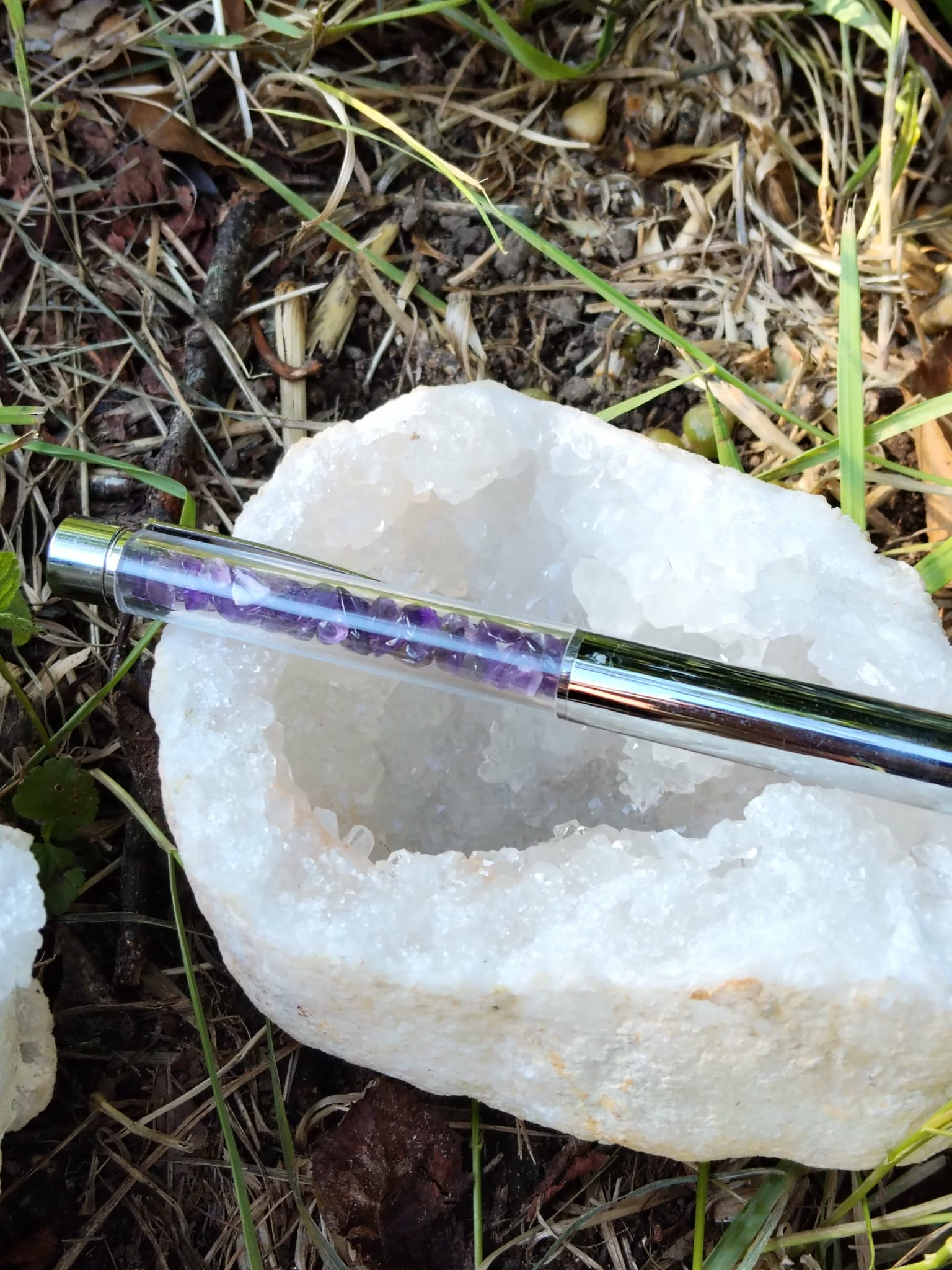 Stylo bille transparent rempli d’éclats d’améthyste, posé sur une grande druse de quartz blanc dans l’herbe, accessoire d’écriture en pierres naturelles pour créativité et calme intérieur.