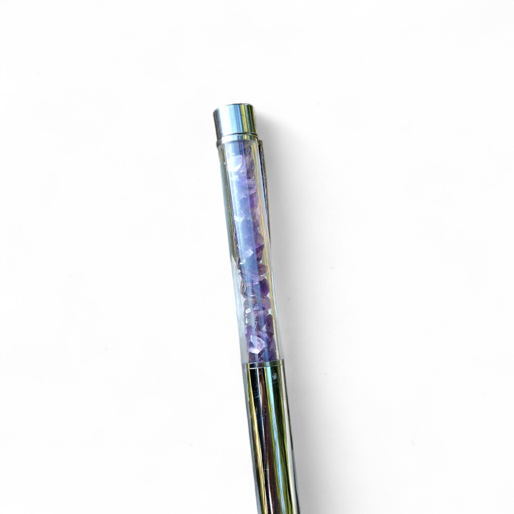 Stylo bille transparent rempli d’éclats d’améthyste violette, photographié verticalement sur fond blanc, accessoire d’écriture en pierres naturelles pour inspiration, écriture intuitive et apaisement du mental.
