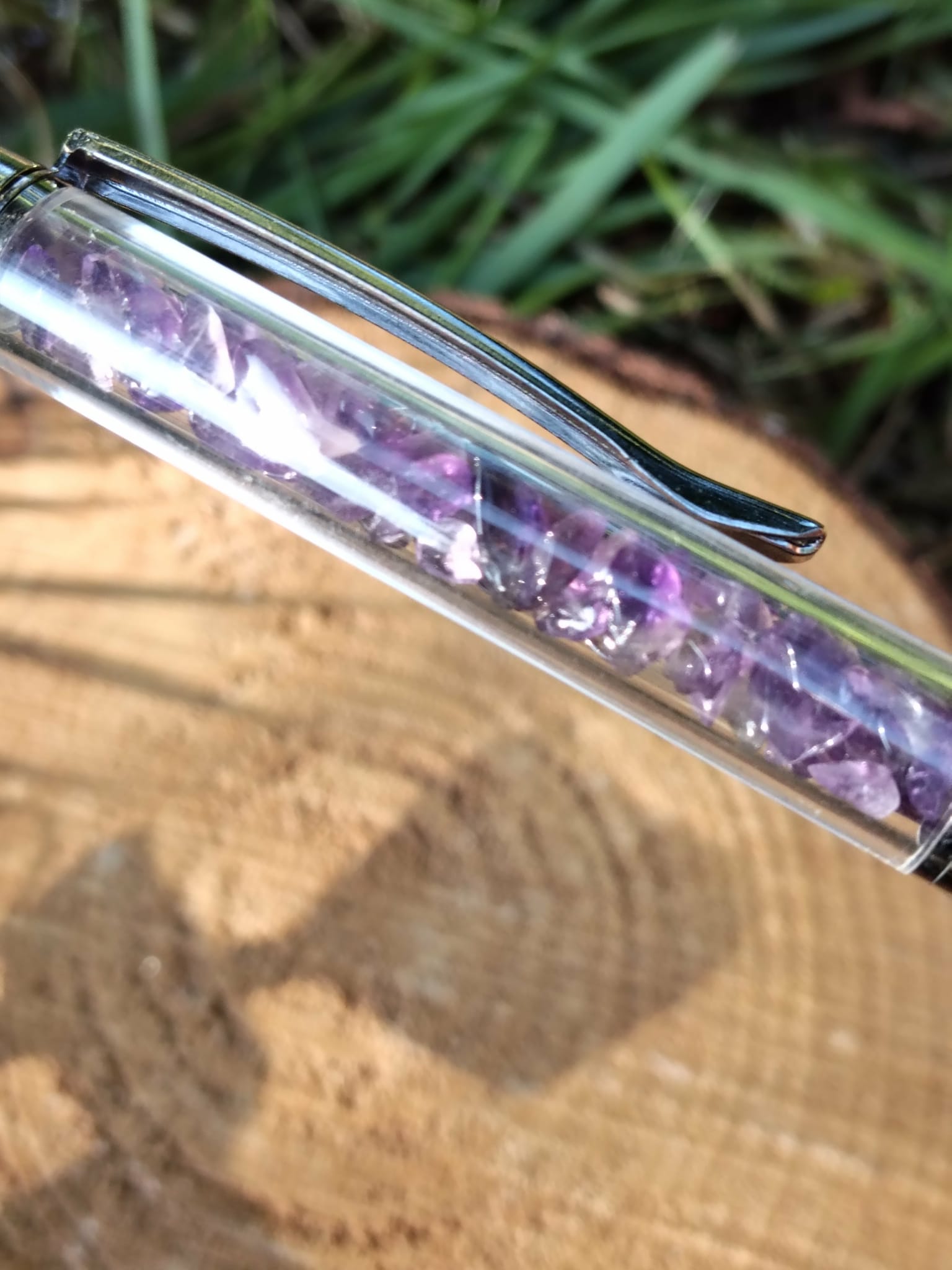 Gros plan sur un stylo bille transparent rempli d’éclats d’améthyste violette, posé en diagonale sur une rondelle de bois avec de l’herbe en arrière-plan, accessoire d’écriture en pierres naturelles favorisant créativité et sérénité.