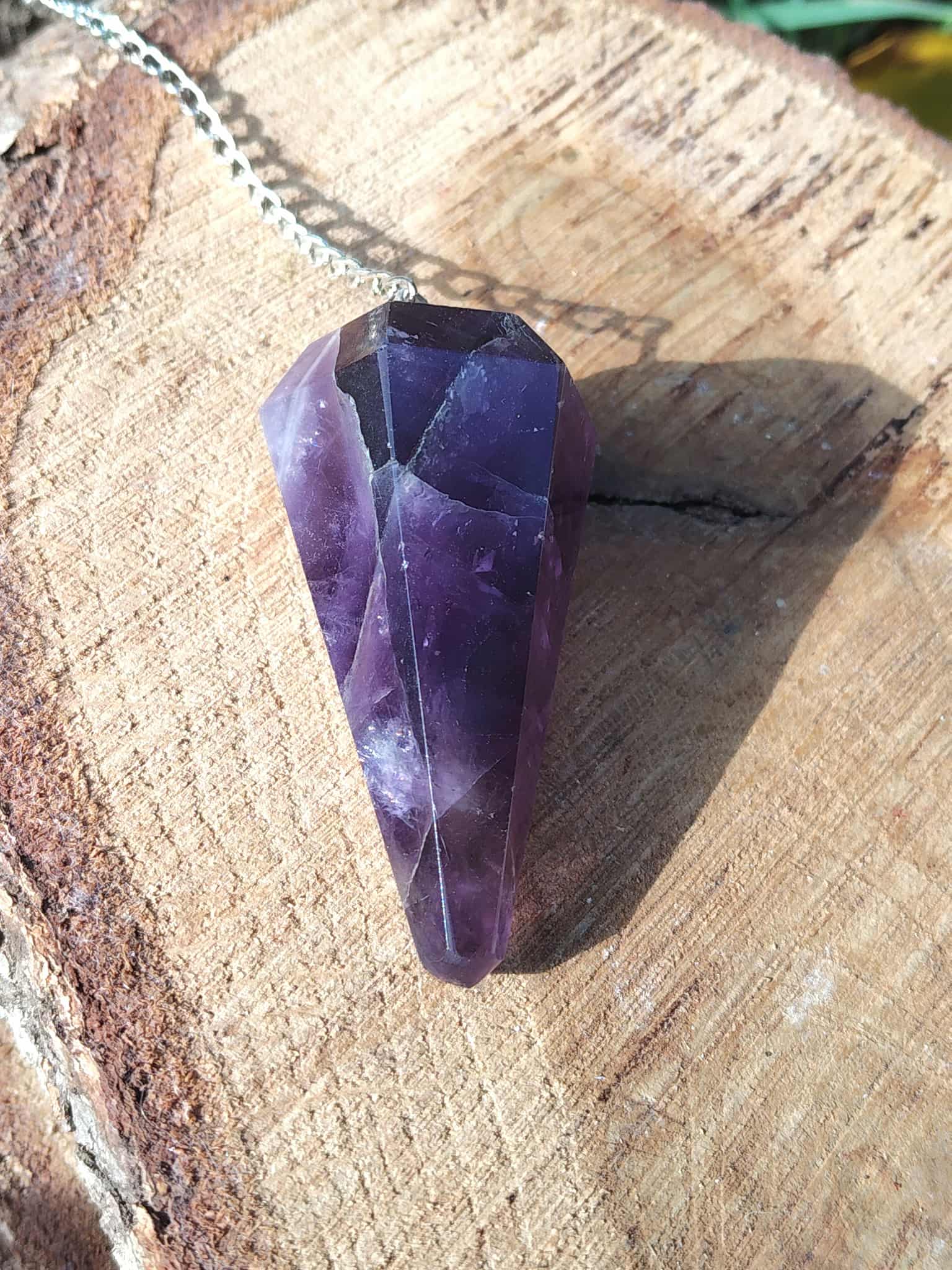 Pendule améthyste en pierre naturelle avec chaîne argentée, pointe facettée posée sur tranche de bois, outil de radiesthésie pour intuition et spiritualité.