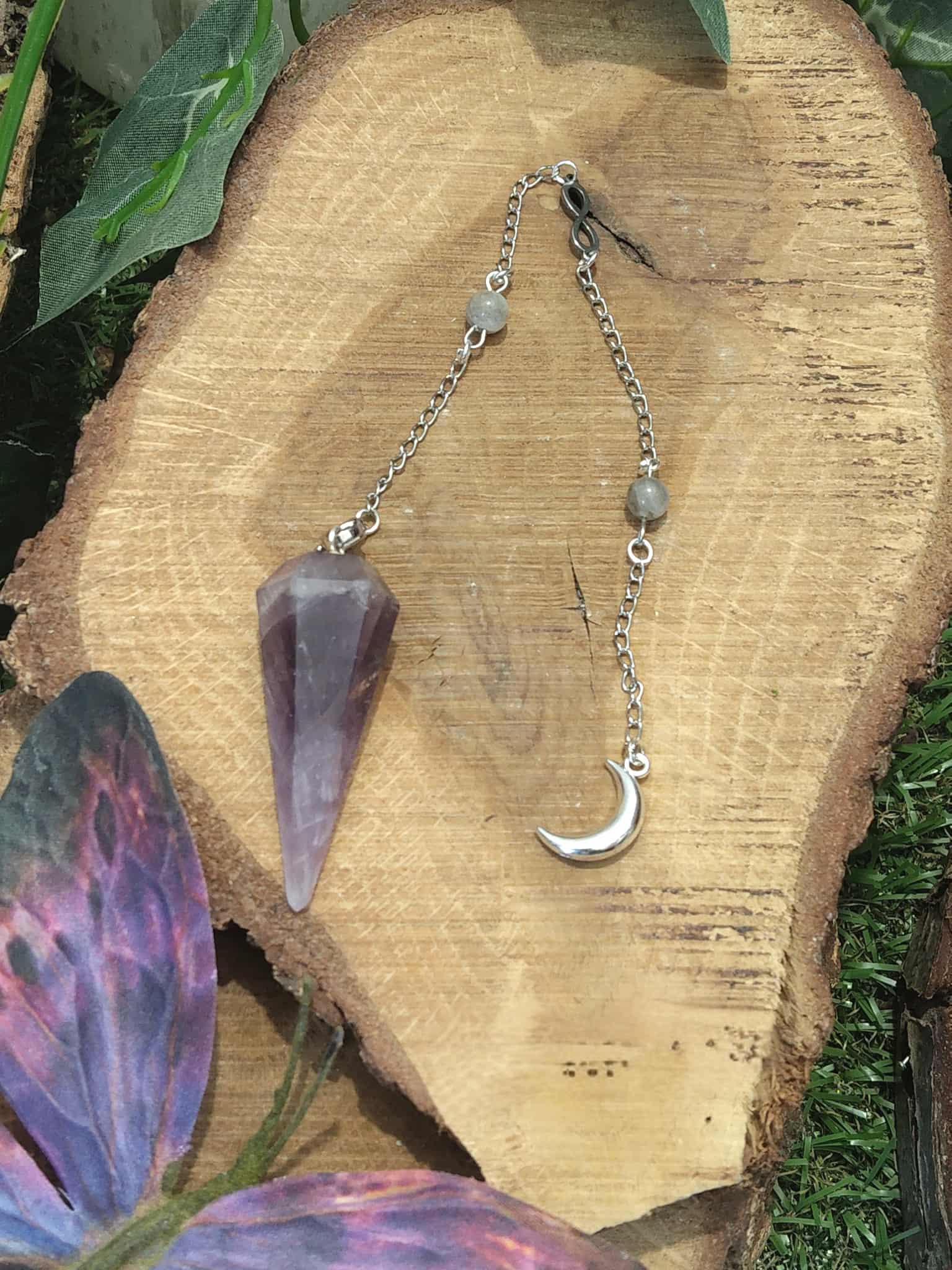 Pendule personnalisé en améthyste naturelle avec chaîne argentée et breloque lune, outil de radiesthésie ésotérique pour intuition et spiritualité, posé sur tranche de bois avec décor végétal.