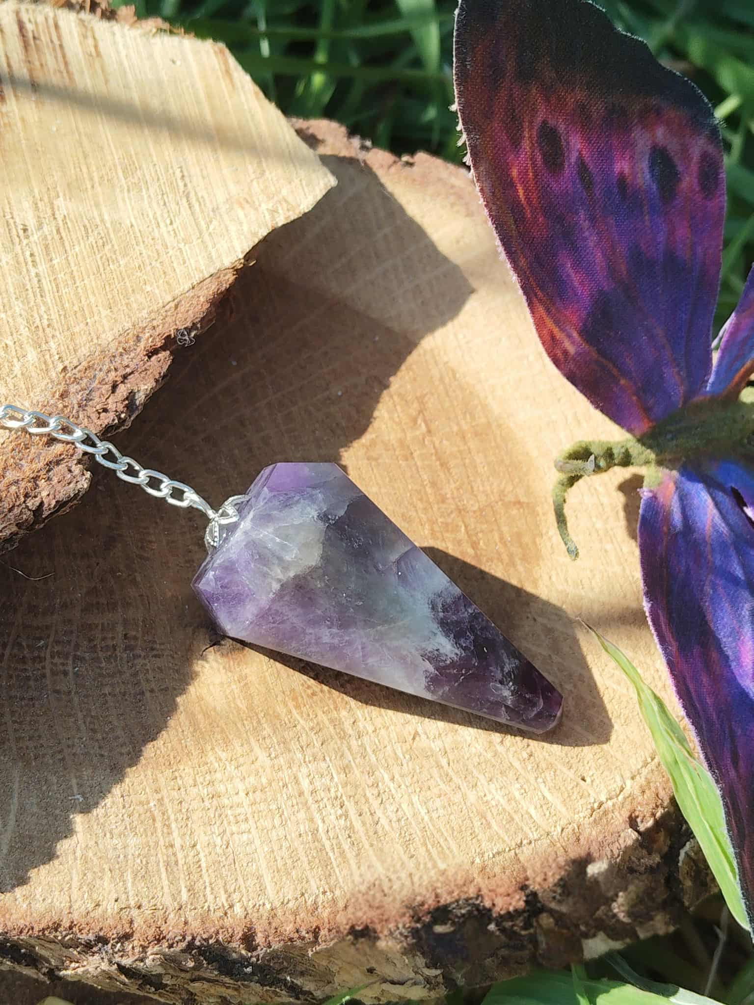 Pendule en améthyste naturelle avec chaîne argentée, posé sur une souche de bois en extérieur avec aile de papillon colorée, outil de radiesthésie ésotérique pour intuition et spiritualité.