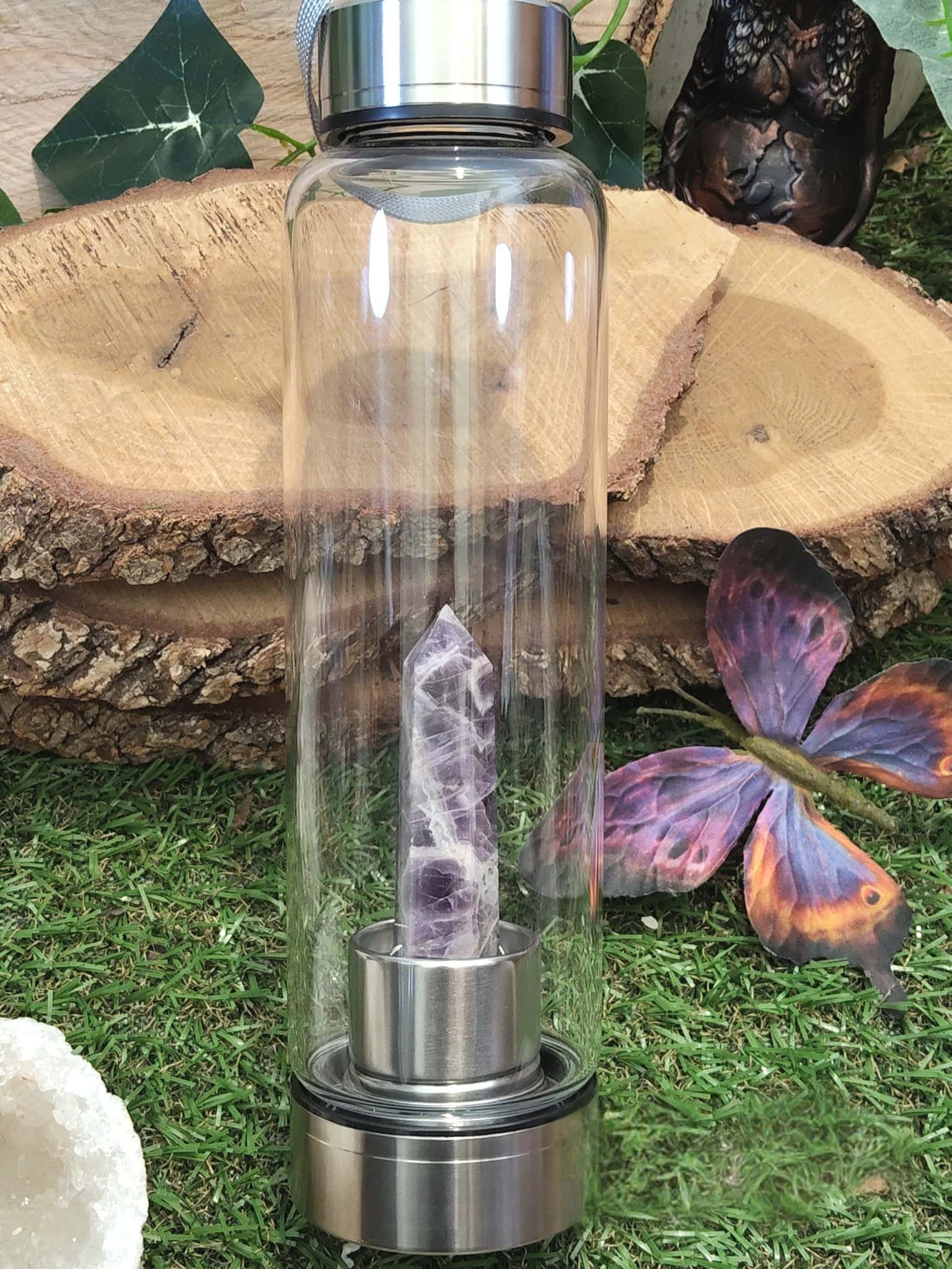 Gourde en verre énergisante améthyste posée sur herbe et rondins de bois, papillon décoratif, objet de lithothérapie pour soutenir le calme intérieur et le rituel d’hydratation.