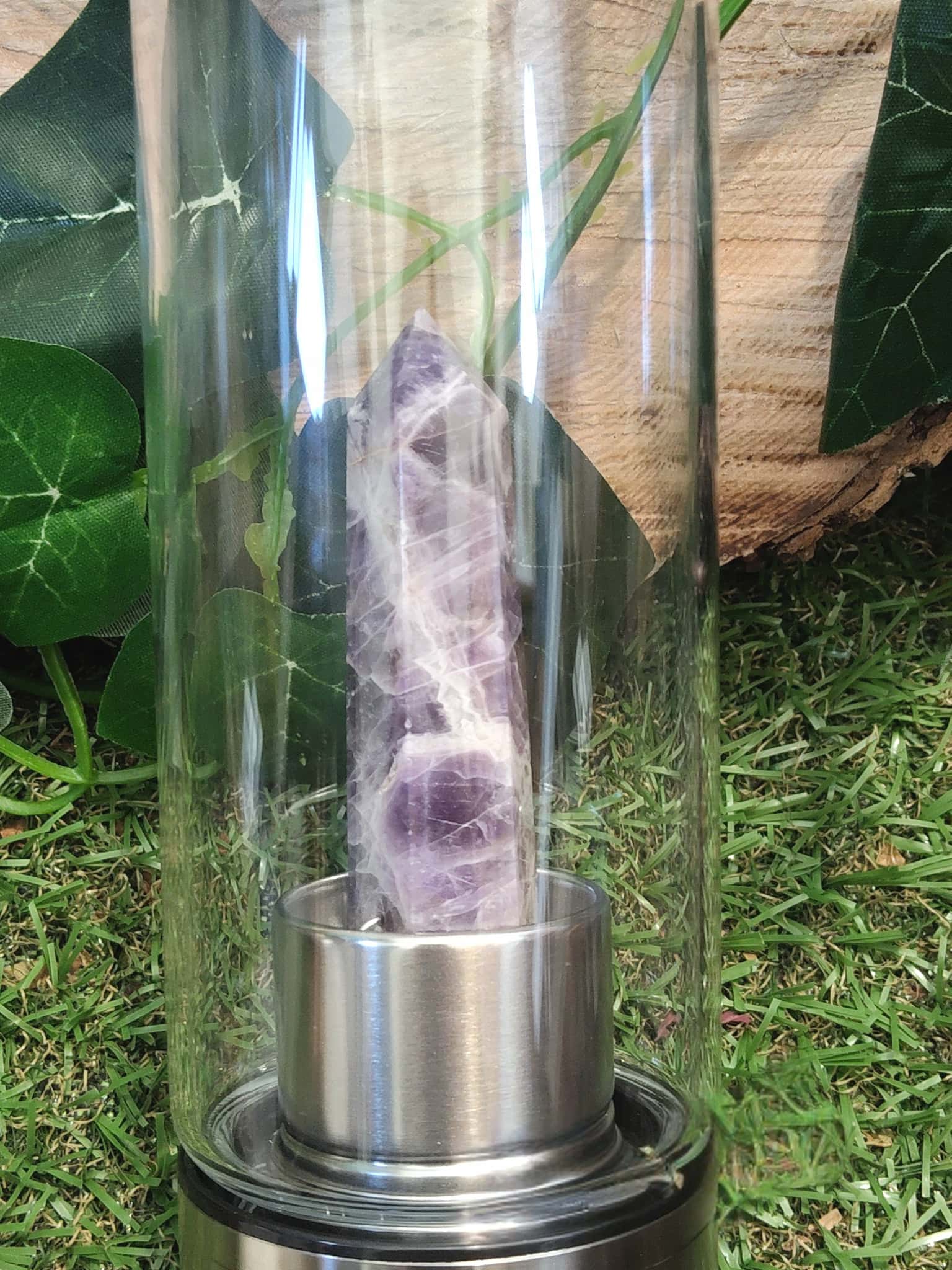 Gros plan sur la pointe d’améthyste naturelle dans la bouteille d’eau en verre énergisante, base inox posée sur l’herbe, accessoire de lithothérapie pour le bien-être.