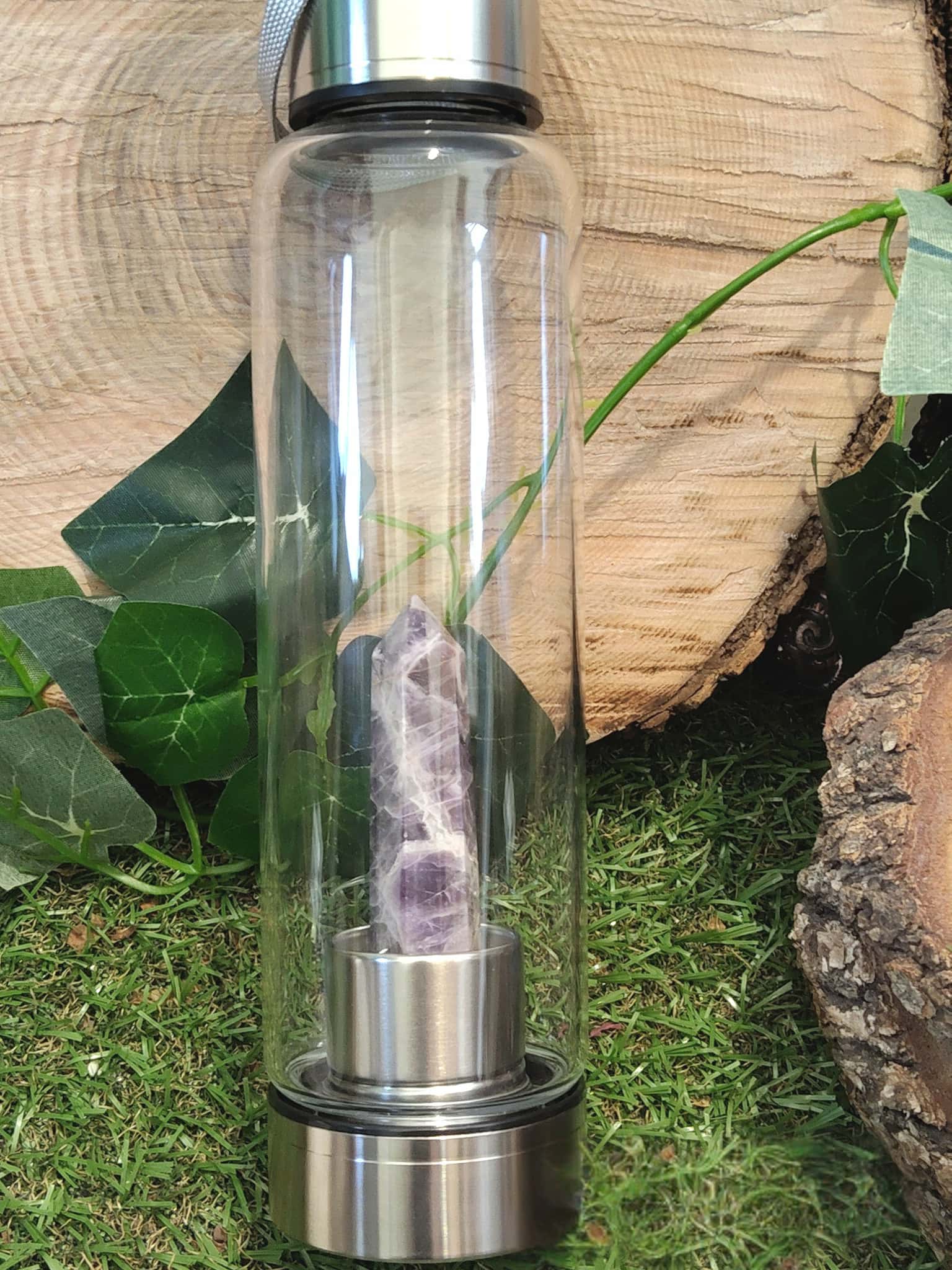 Bouteille d’eau en verre énergisante avec pointe d’améthyste naturelle, 500 ml, posée sur un décor végétal et bois brut, accessoire de lithothérapie pour l’hydratation consciente, le bien-être énergétique et l’apaisement.