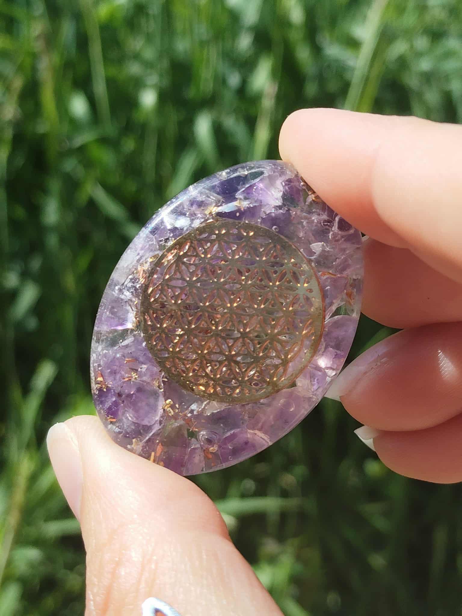 Petit galet orgonite améthyste avec fleur de vie dorée tenu entre les doigts devant un fond de nature, galet énergétique de lithothérapie pour harmonisation des énergies, protection, apaisement mental et émotionnel