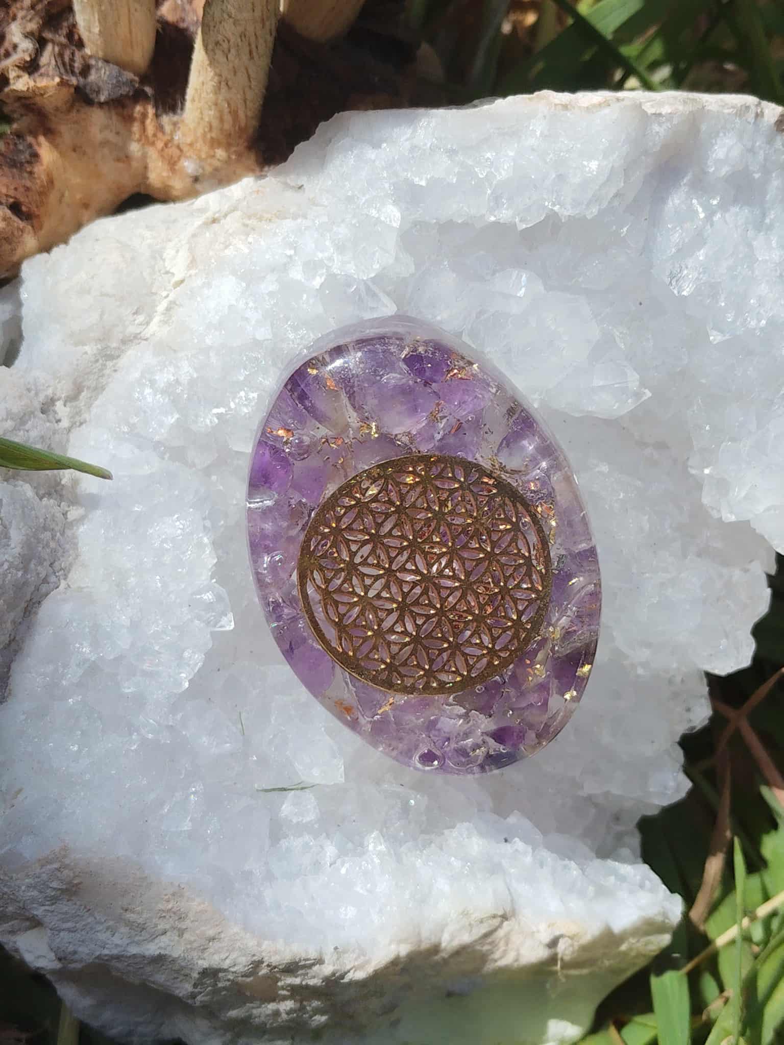Petit galet orgonite améthyste avec fleur de vie dorée posé sur une grande géode de cristal blanc en extérieur, objet de lithothérapie pour protection énergétique, harmonisation des énergies et décoration spirituelle