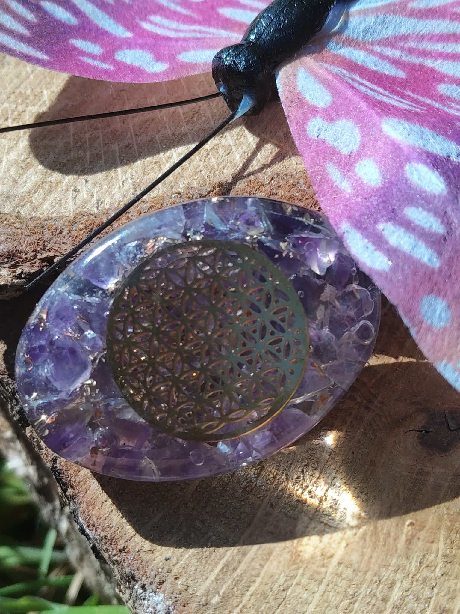 Petit galet énergétique en orgonite améthyste avec symbole fleur de vie, posé sur une planche de bois avec papillon décoratif rose et bleu, objet de lithothérapie pour protection, harmonisation des énergies et décoration spirituelle