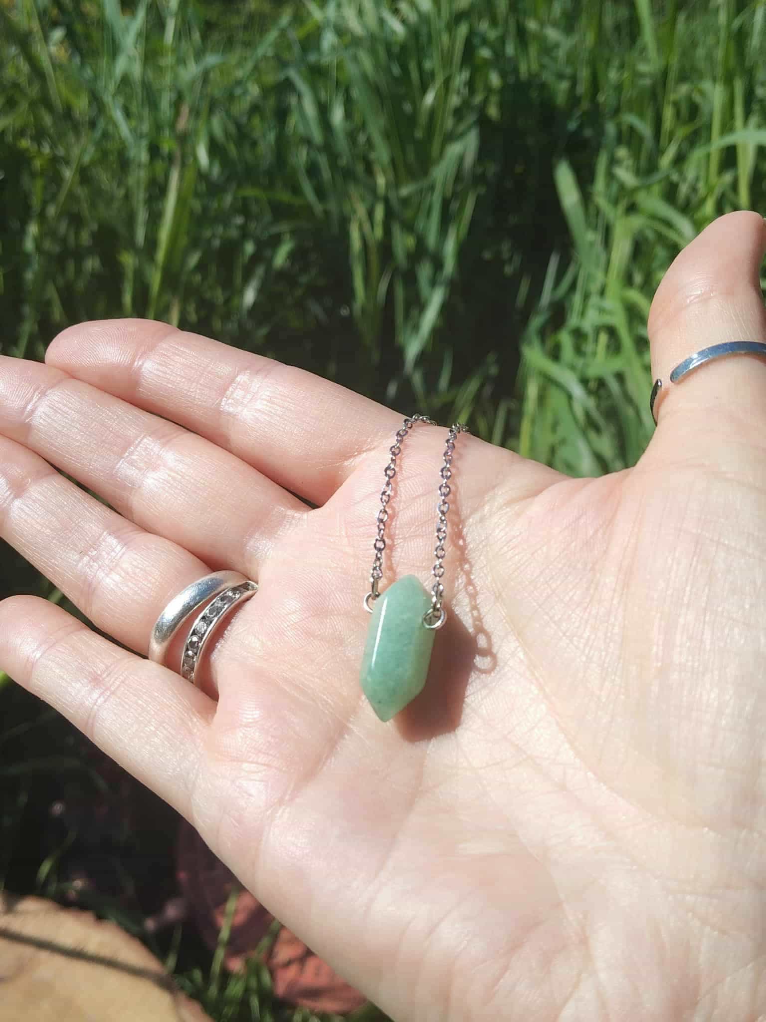 Pendentif en pointe d’aventurine verte tenu dans la main sur fond d’herbe, collier en pierre naturelle associé à la confiance douce, au calme des pensées et à l’équilibre émotionnel.
