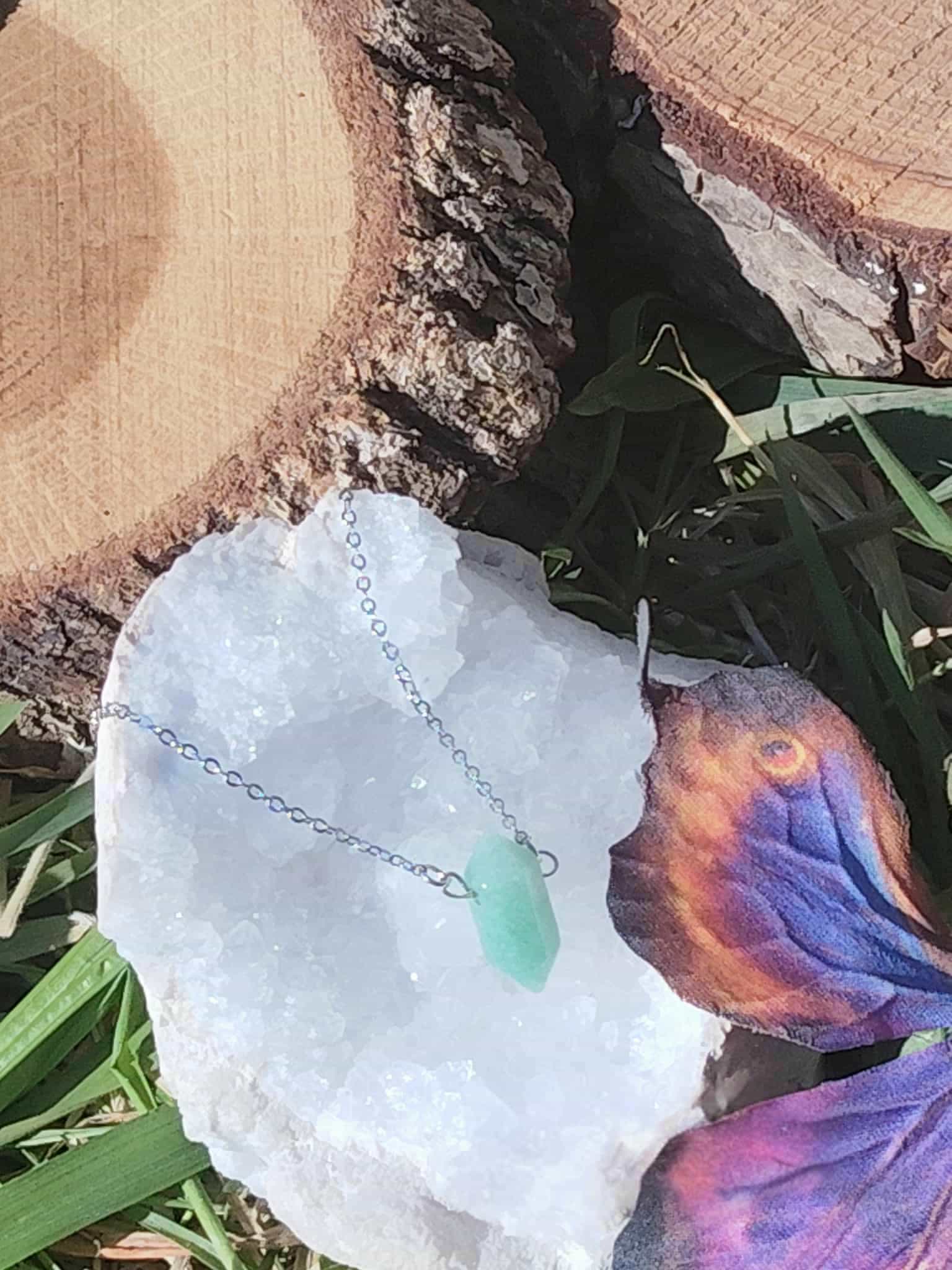 Pendentif pointe aventurine verte sur chaîne fine, posé sur géode blanche avec fleurs et rondins de bois en extérieur, bijou pierre naturelle associé à l’ouverture du cœur, à la fraîcheur émotionnelle et au calme.