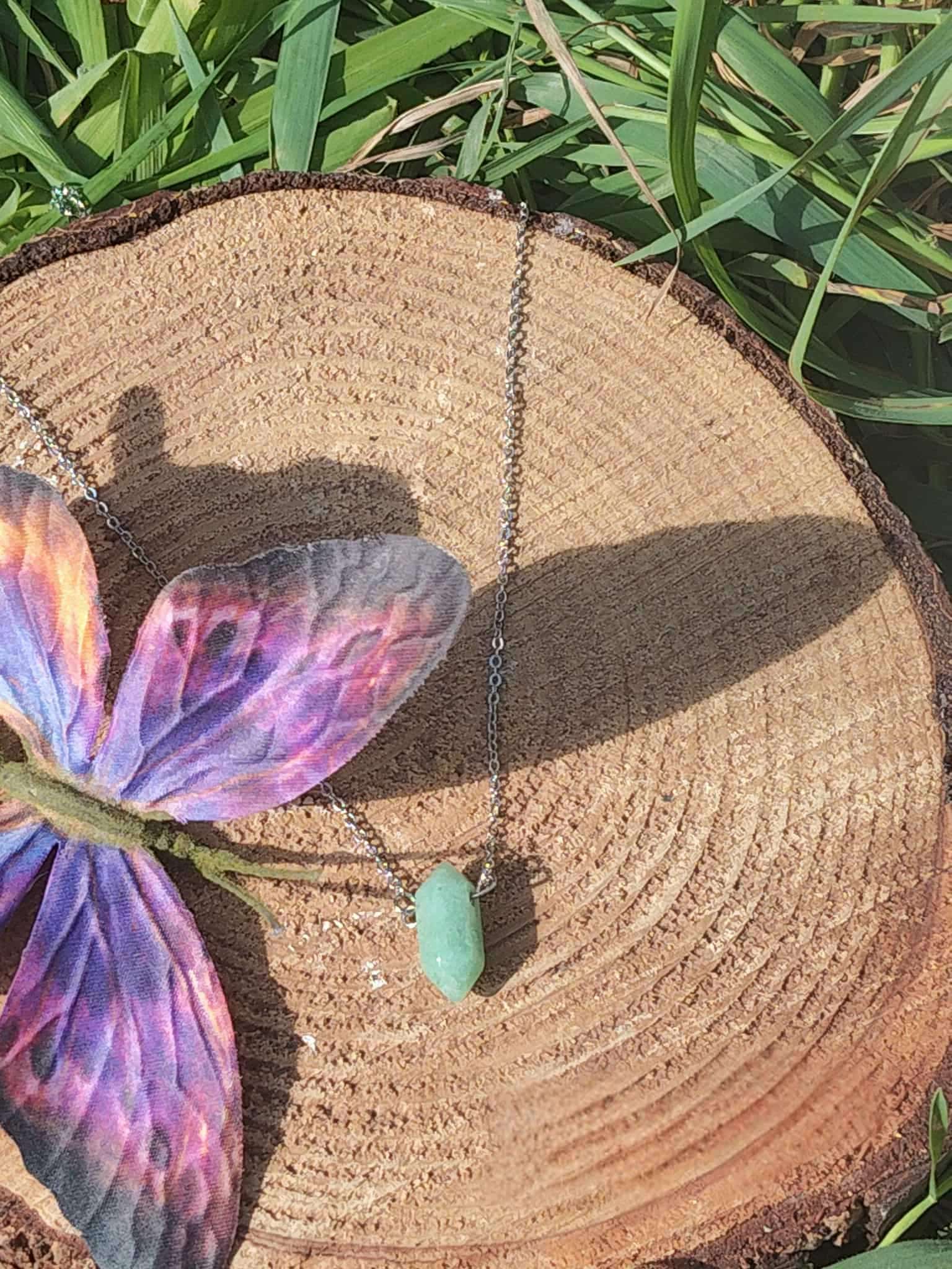 Collier avec pendentif en pointe d’aventurine verte posé sur rondin de bois, décor papillon multicolore et herbe, bijou pierre naturelle relié à la légèreté, à l’apaisement affectif et à la sensibilité du cœur.