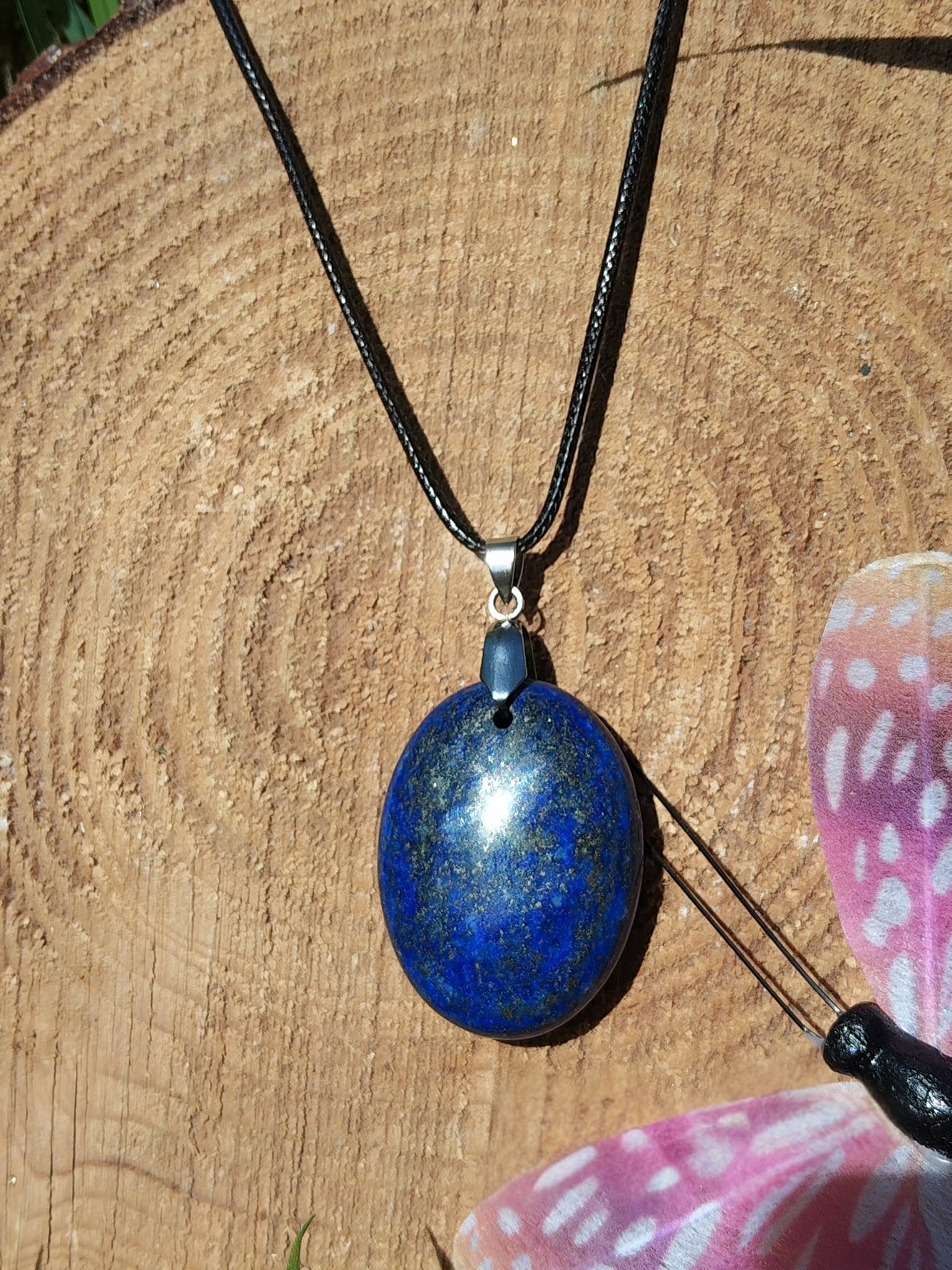 Pendentif rond en lapis lazuli bleu sur cordon noir, posé sur bois avec papillon rose, collier pierre naturelle évoquant l’assurance, l’écoute de soi et la paix intérieure.