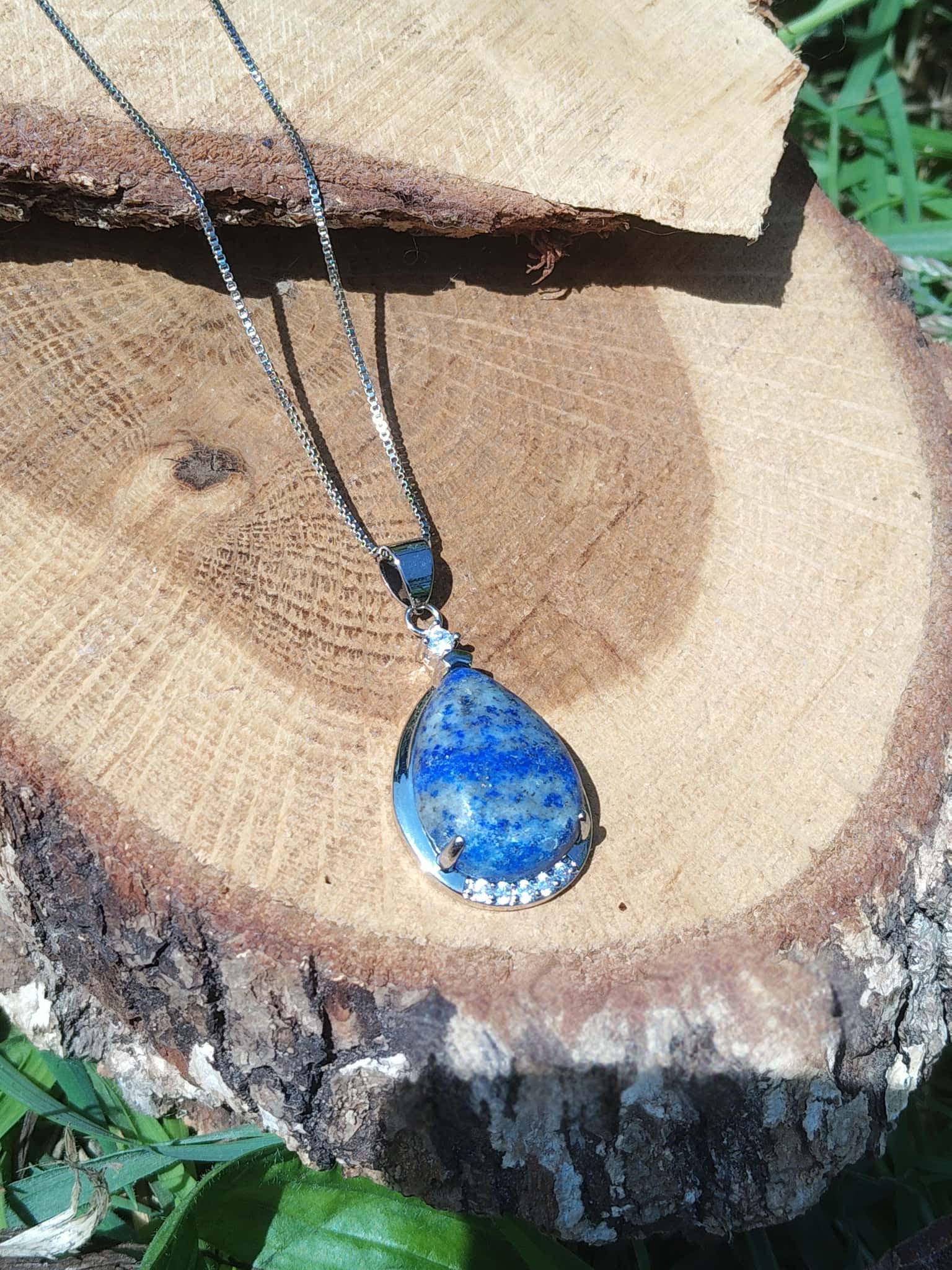 Collier avec pendentif goutte en lapis lazuli bleu posé sur rondin de bois, bijou en pierre naturelle associé à la maîtrise de soi, à la lucidité et à l’équilibre émotionnel.