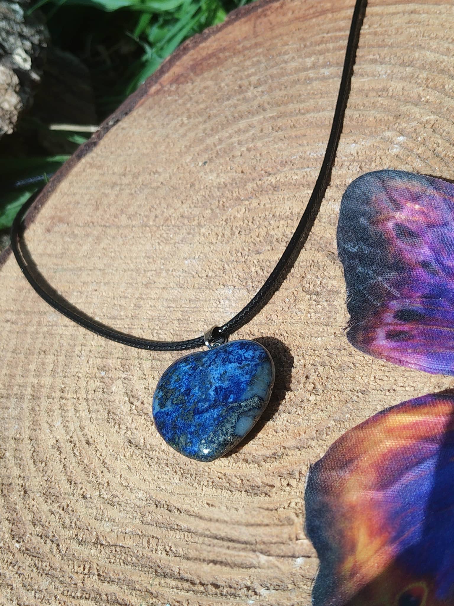 Collier cœur en lapis lazuli bleu sur rondin de bois avec ailes colorées, bijou pierre naturelle associé à la sensibilité, à la lucidité et à la douceur émotionnelle.
