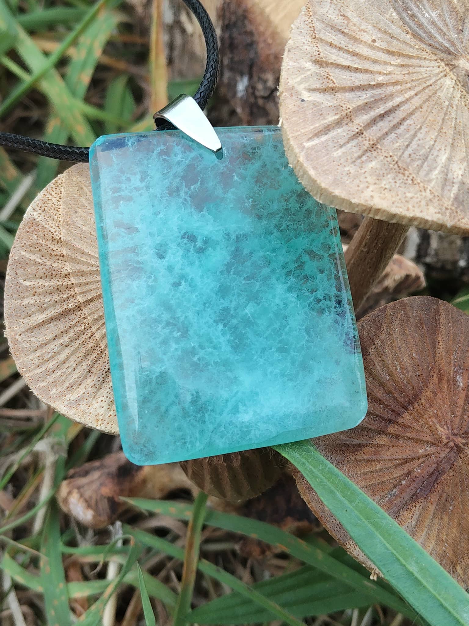 Gros plan sur pendentif rectangulaire en fluorite verte posé sur rondins de bois dans l’herbe, collier en pierre naturelle relié à la sérénité, à l’ordre des pensées et à la présence à soi.
