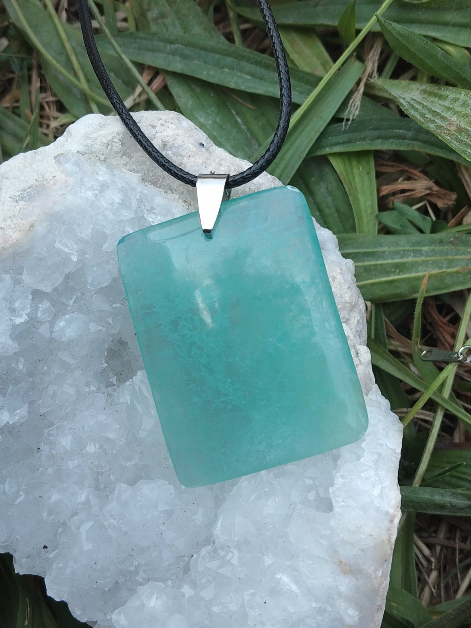 Pendentif fluorite verte rectangulaire sur cordon noir posé sur géode blanche dans l’herbe, pierre naturelle associée à l’apaisement, au lâcher-prise et à l’équilibre intérieur.