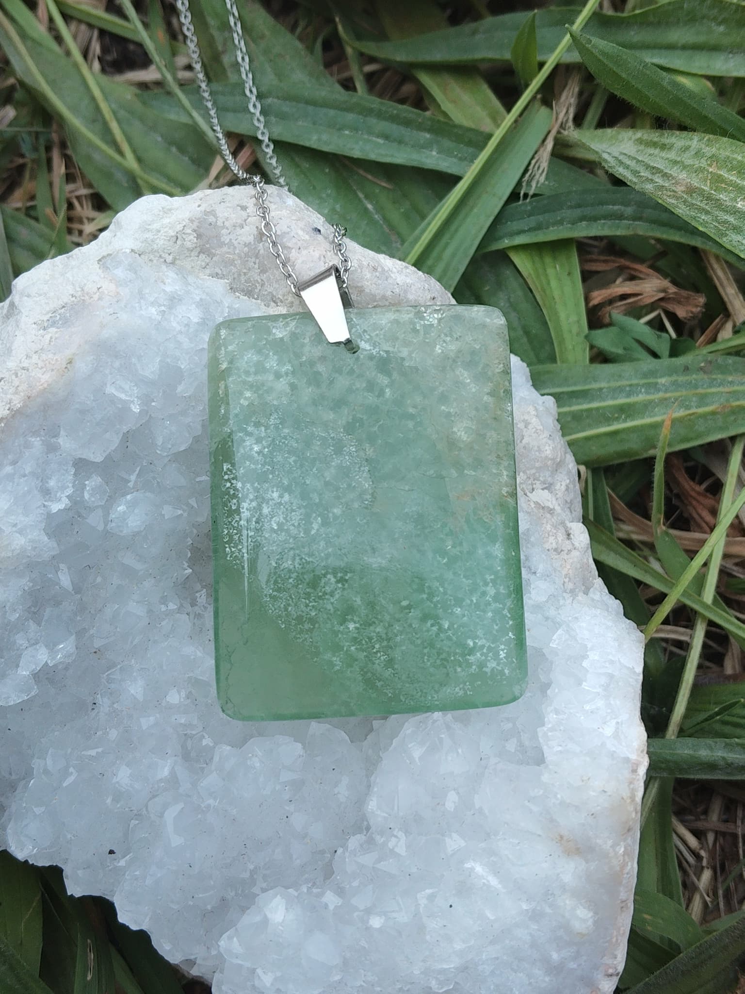 Pendentif rectangulaire en fluorite verte sur chaîne argentée posé sur géode blanche dans l’herbe, bijou naturel évoquant la finesse intuitive, la respiration émotionnelle et le calme mental.