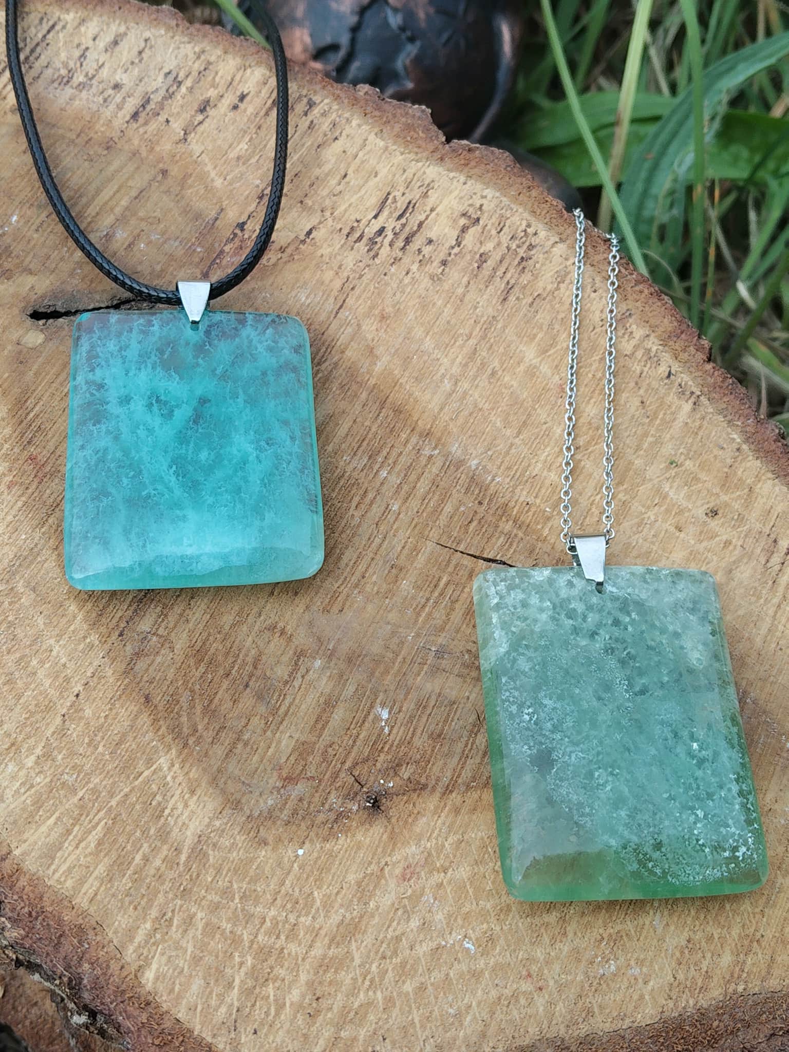 Deux pendentifs rectangulaires en fluorite verte sur cordon noir et chaîne argentée, posés sur une tranche de bois, bijoux en pierre naturelle associés à la tendresse, à la clarté et à l’équilibre émotionnel.