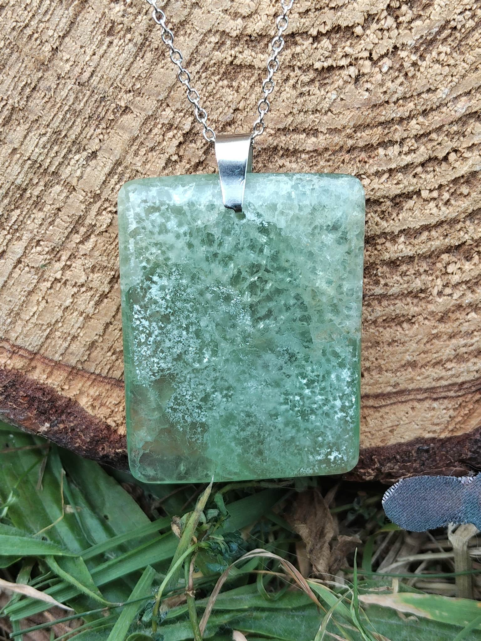Pendentif rectangulaire en fluorite verte sur chaîne argentée, posé devant un rondin de bois et de l’herbe, pierre naturelle liée à la légèreté intérieure, à la justesse émotionnelle et au recentrage.