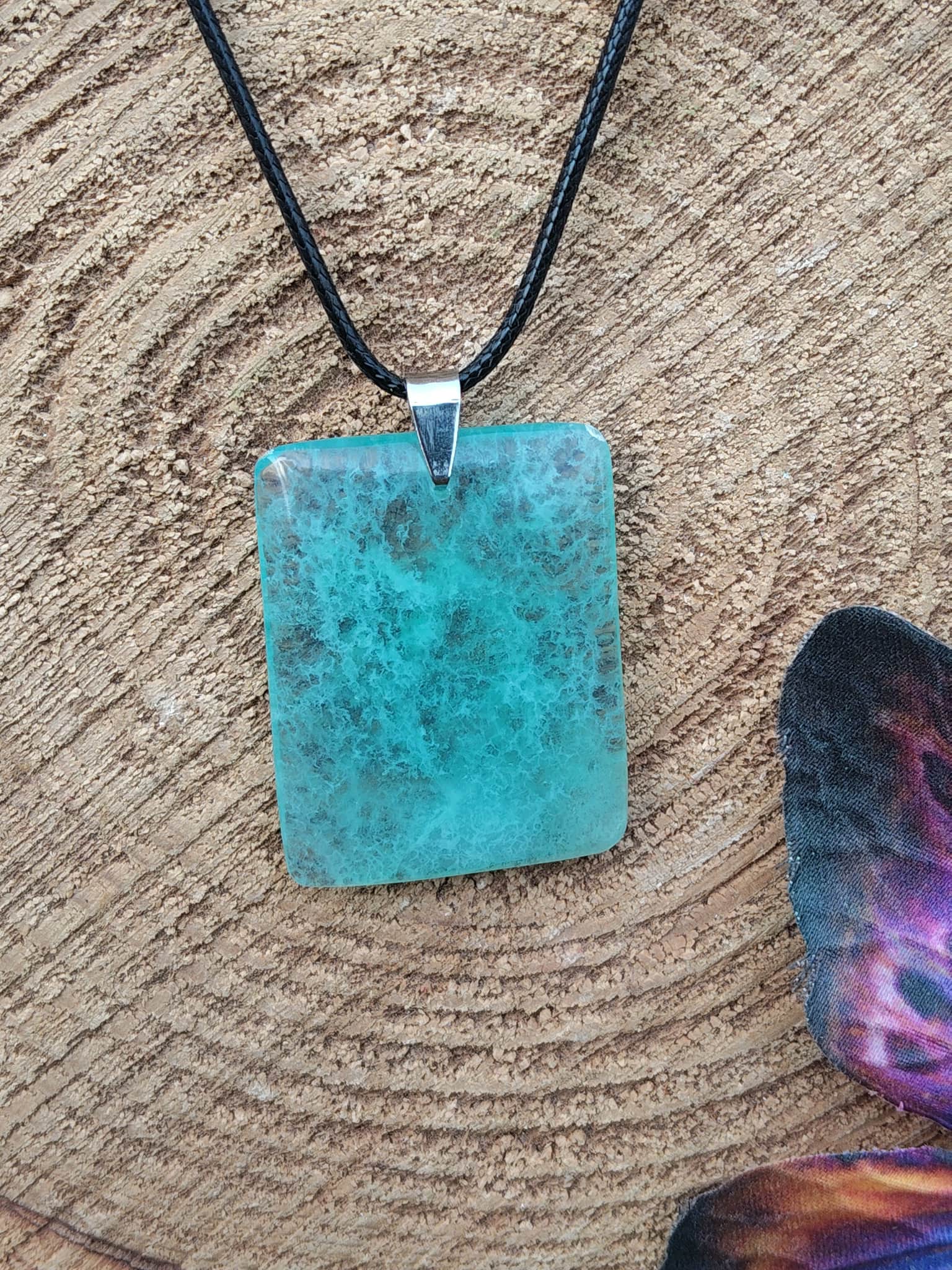 Pendentif rectangulaire en fluorite verte sur cordon noir, posé sur rondin de bois avec décor ésotérique, collier en pierre naturelle évoquant la concentration paisible, le recul et l’harmonie émotionnelle.