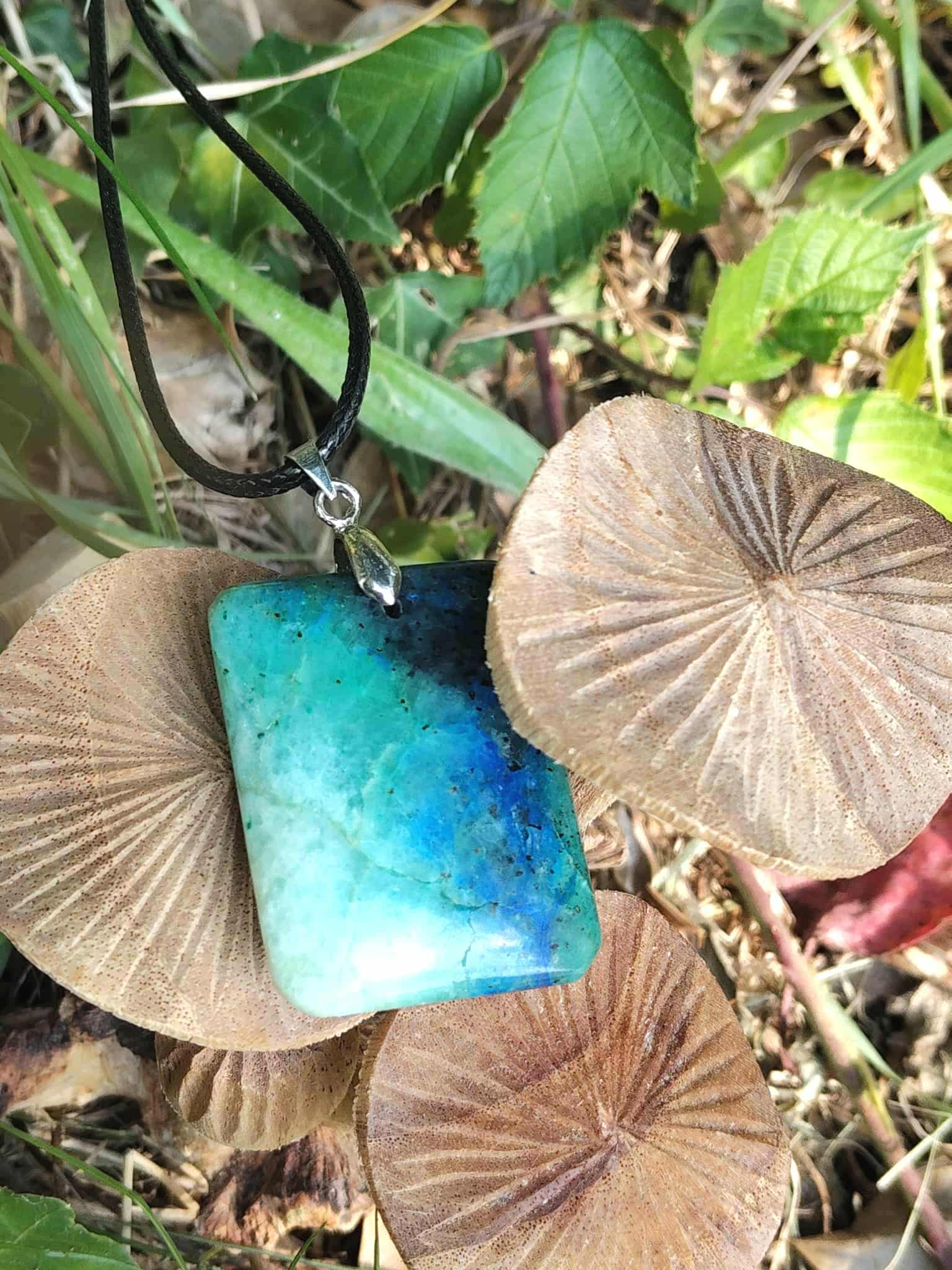Pendentif azurite chrysocolle bleu vert rectangulaire sur cordon noir, posé parmi des champignons et feuilles en sous-bois, pierre naturelle évoquant la tranquillité, la présence à soi et la communication apaisée.