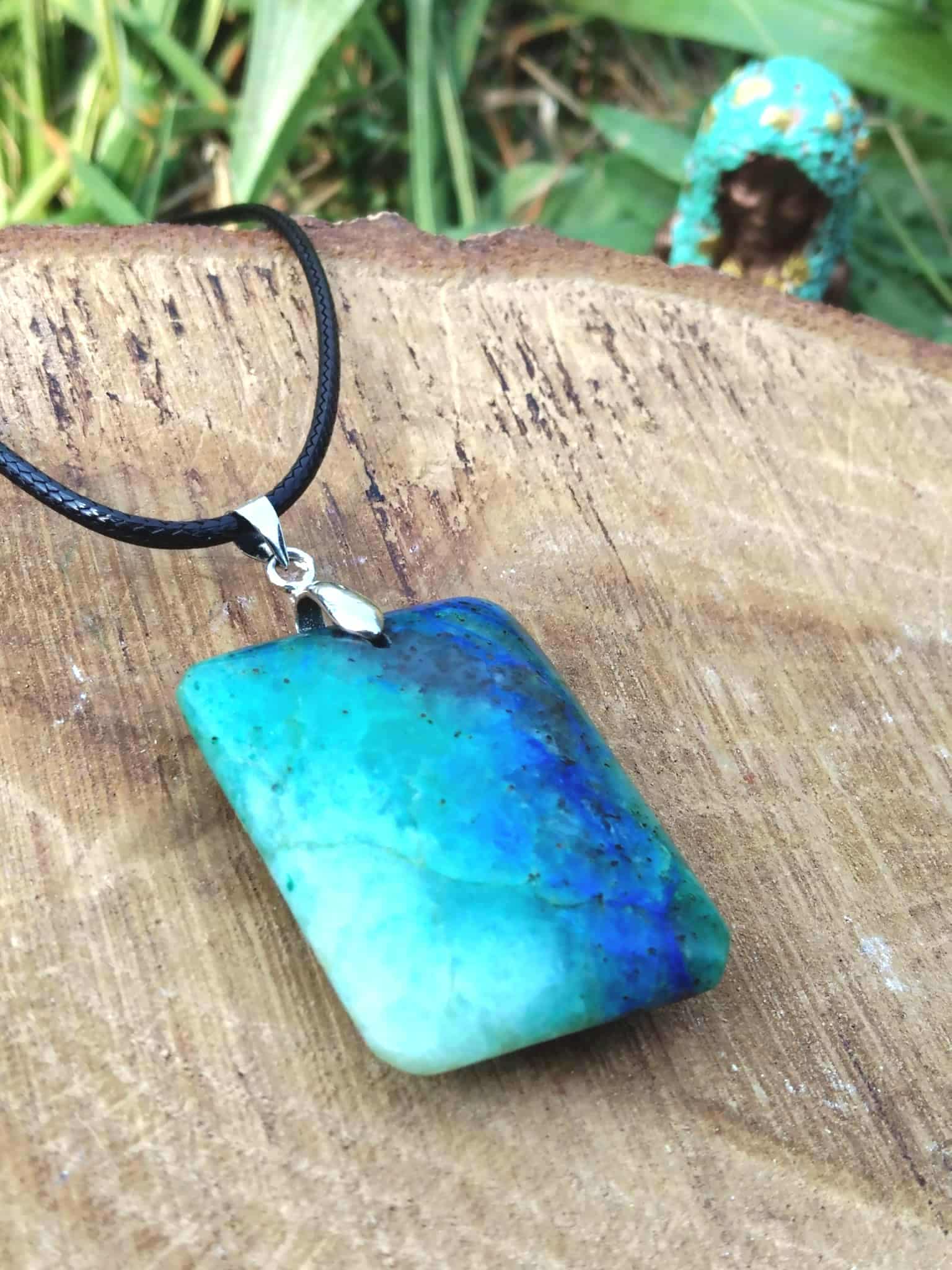 Pendentif azurite chrysocolle bleu vert sur cordon noir, posé sur bois avec herbes en arrière-plan, pierre naturelle associée à la parole fluide, à l’équilibre affectif et à la paix intérieure.