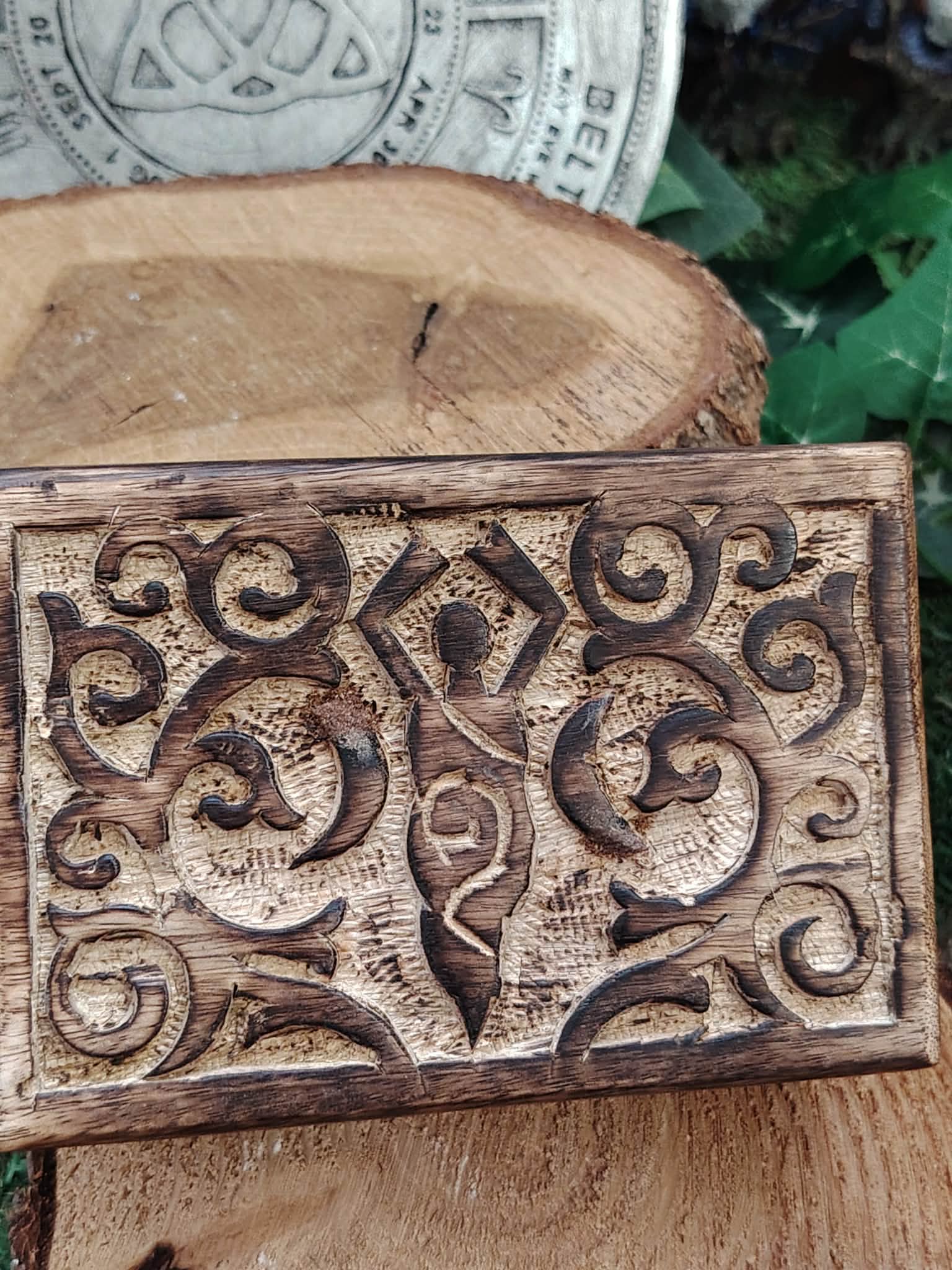 Boîte à bijoux en bois sculpté motif déesse, posée sur une tranche de bois, avec une grande rondelle de bois, un disque décoratif gris et du feuillage vert en arrière-plan.