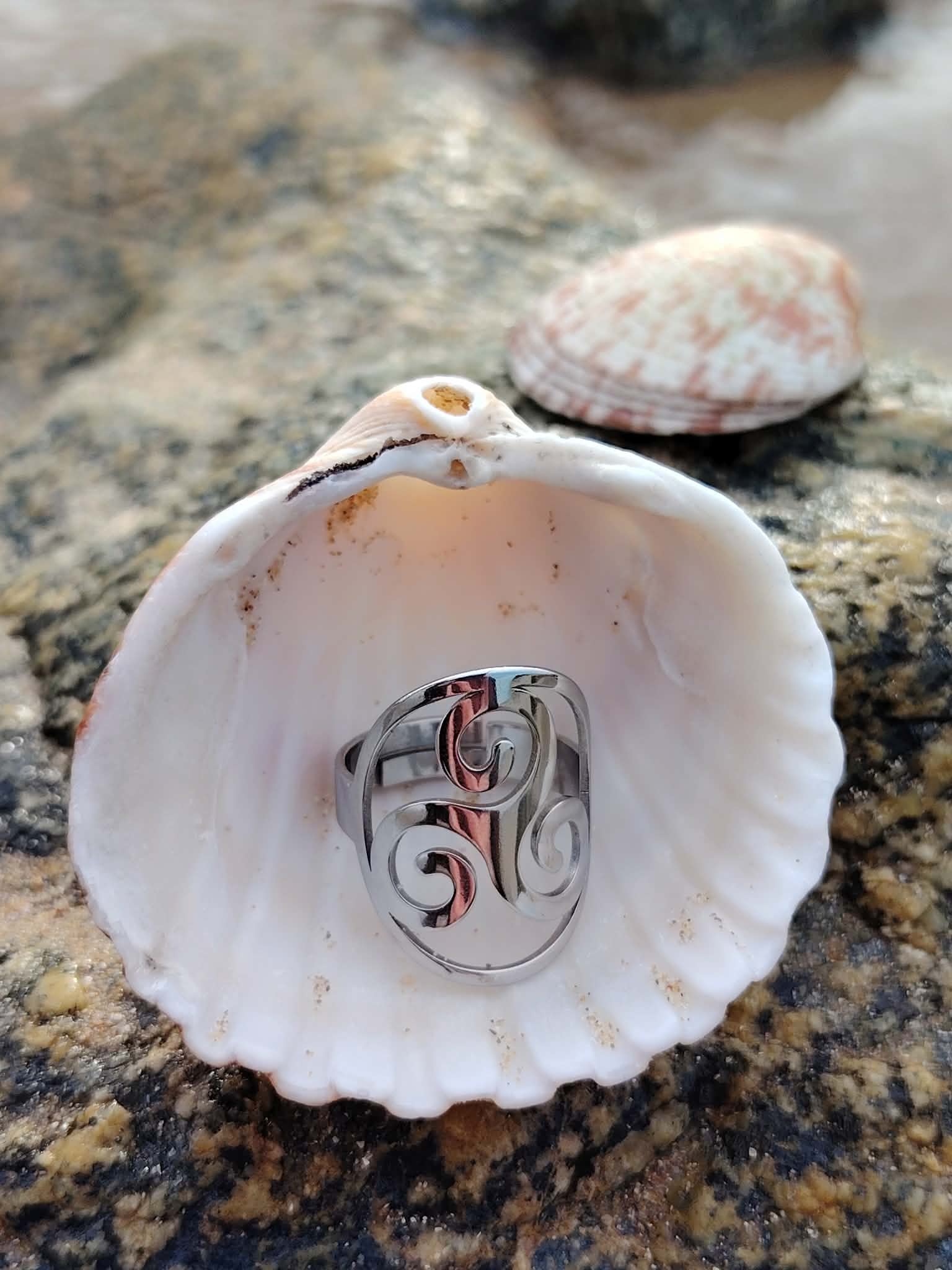 Bague Triskell réglable en acier inoxydable, symbole celtique de protection et d’harmonie, présentée dans un coquillage ouvert sur un rocher en bord de mer en Bretagne bijou Magic Gaïa