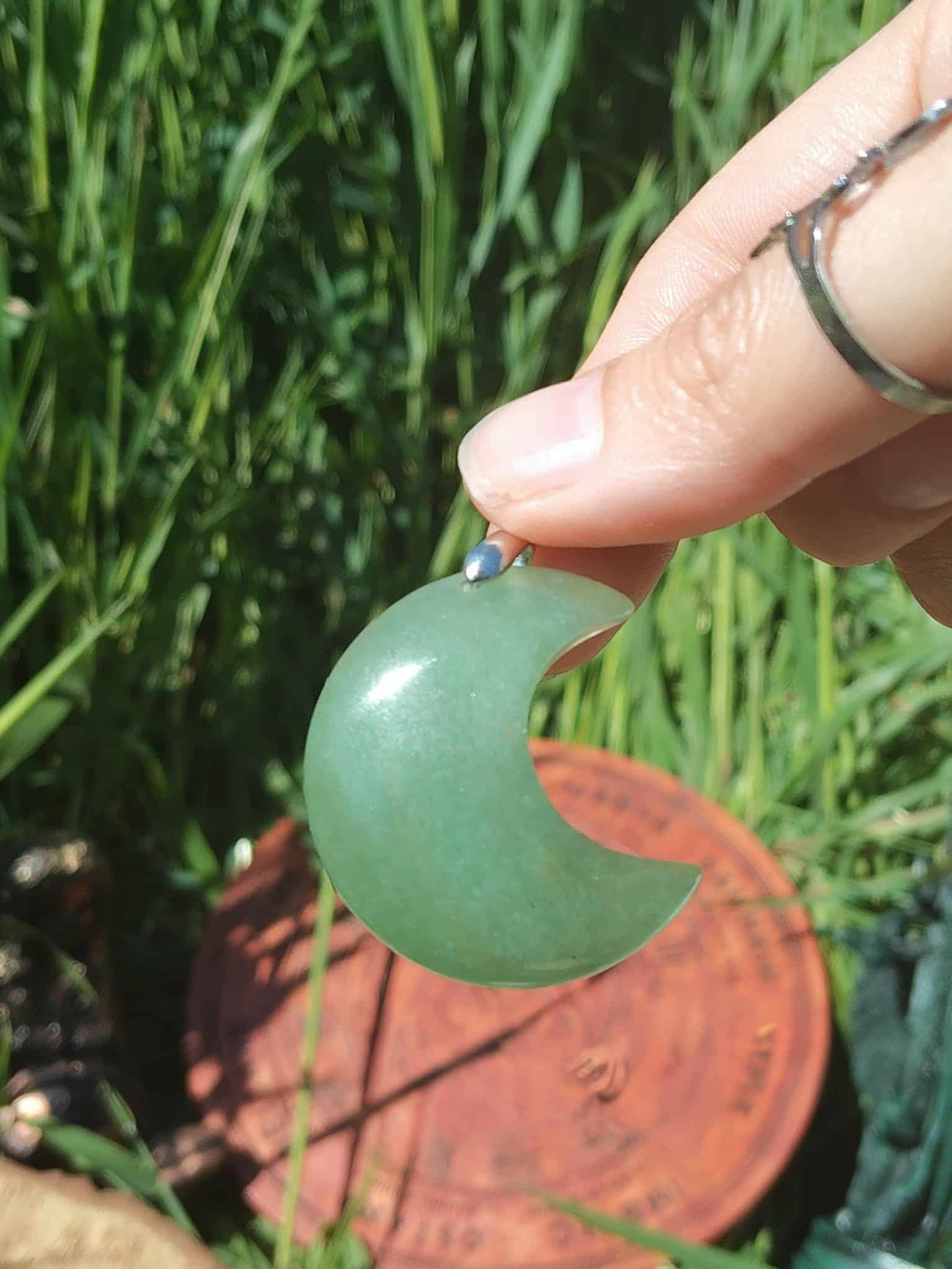 Pendentif lune en aventurine verte tenu entre les doigts devant l’herbe, bijou en pierre naturelle évoquant le lâcher-prise, la tendresse et l’équilibre affectif.
