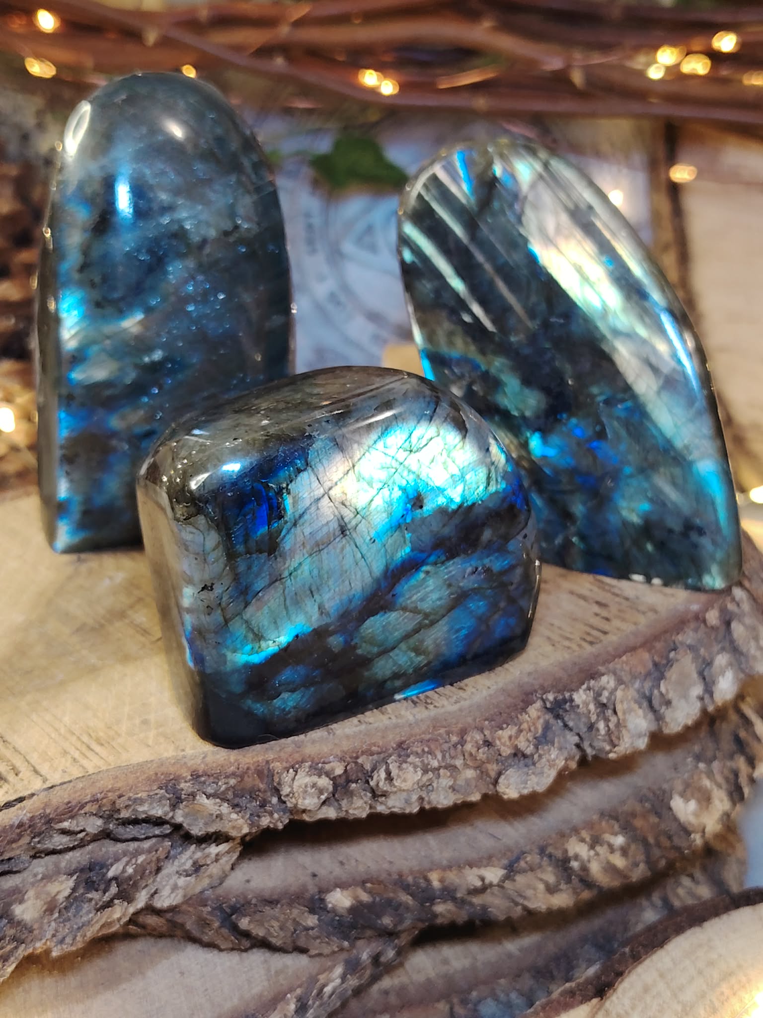 Trio de labradorites forme libre posé sur un rondin de bois brut avec guirlande lumineuse, pierres naturelles évoquant la protection, la force calme et l’enracinement intérieur.