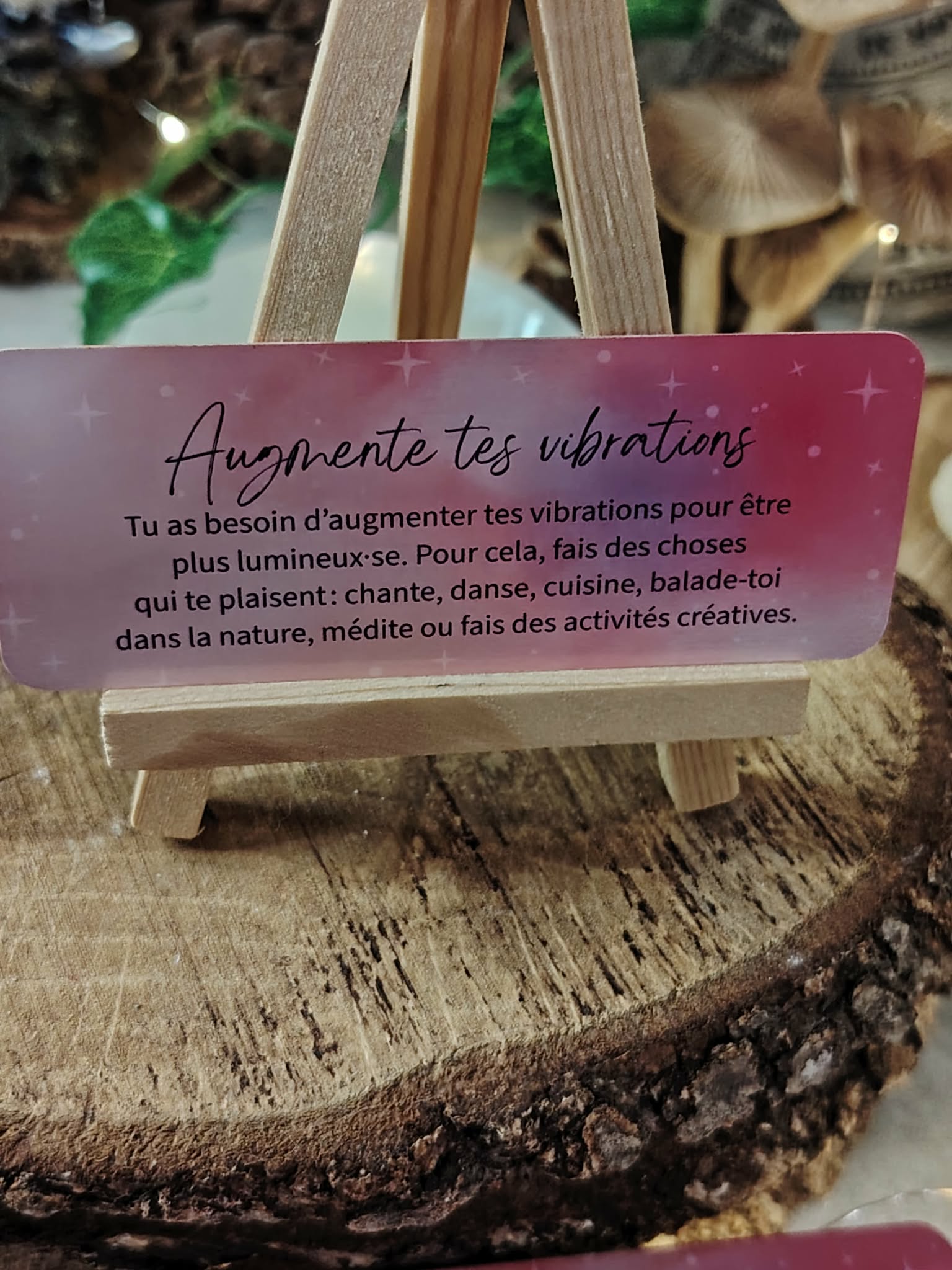 Carte “Augmente tes vibrations” du Petit Oracle des Sœurcières (L’Atelier des Sœurcières) posée sur chevalet en bois, ambiance naturelle sur rondin, décor chaleureux et spirituel