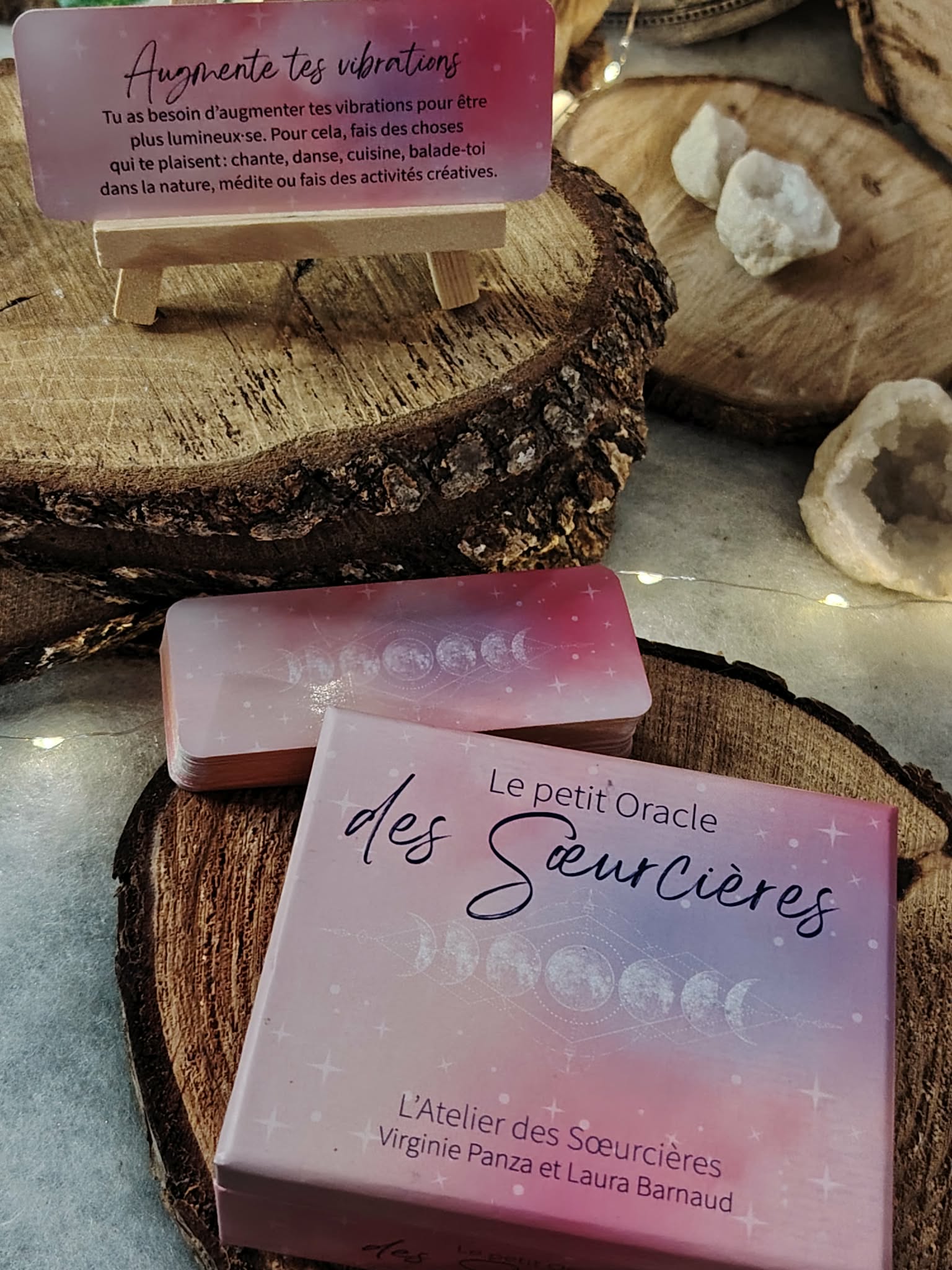 Le Petit Oracle des Sœurcières (L’Atelier des Sœurcières) : boîte rose et cartes oracle en français posées sur rondins de bois, avec pierres de quartz blanc et guirlande lumineuse – Magic Gaïa