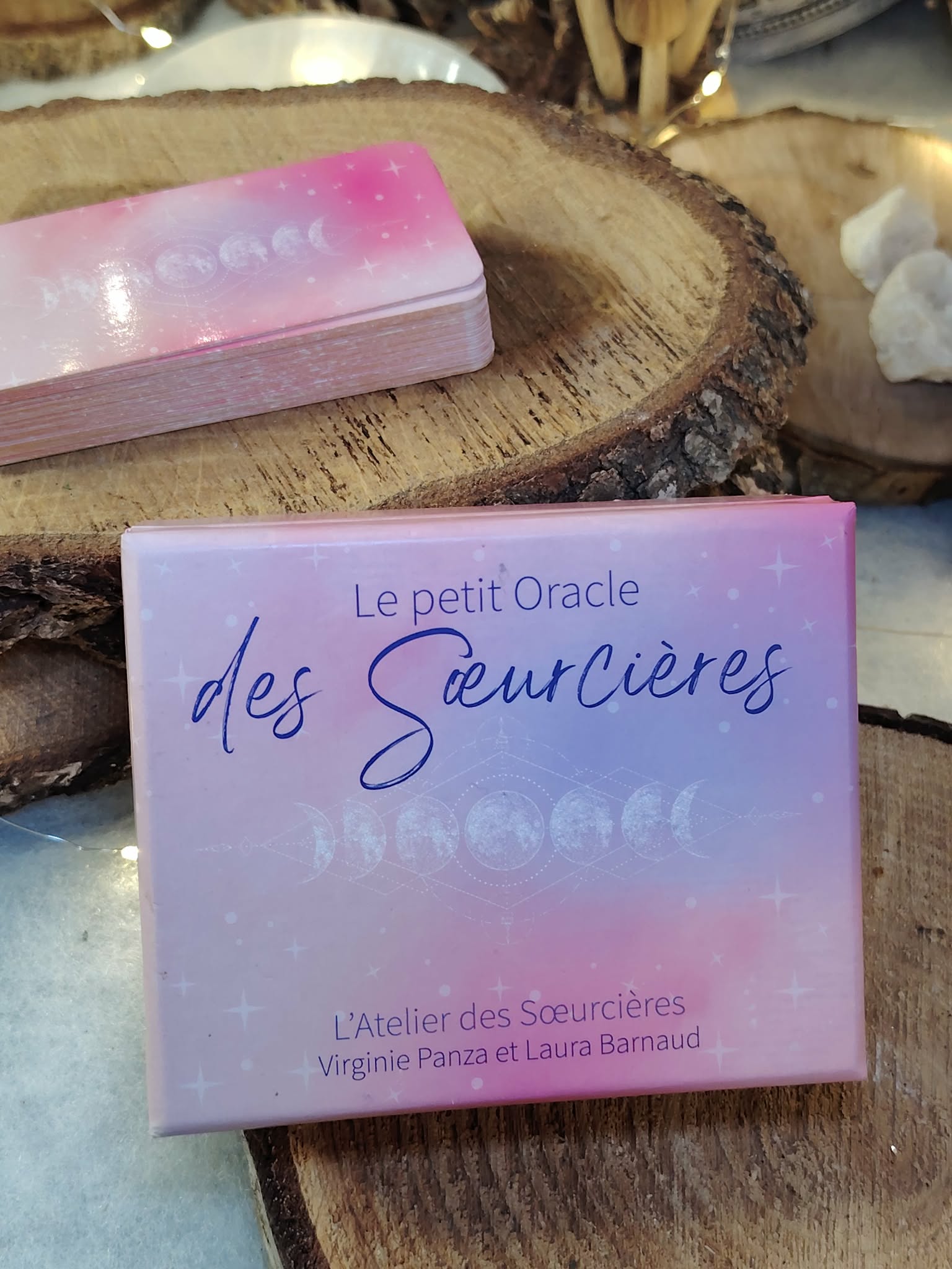 Boîte Le Petit Oracle des Sœurcières (L’Atelier des Sœurcières) en français, coffret rose/lilas posé sur rondin de bois, décor ésotérique avec pierres de quartz blanc et ambiance chaleureuse