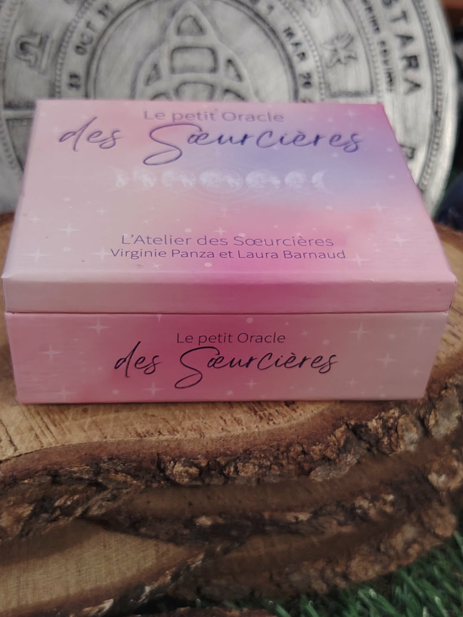 Coffret Le Petit Oracle des Sœurcières (L’Atelier des Sœurcières) : boîte rose/lilas vue de côté sur rondin de bois, décor ésotérique avec fond mandala et ambiance chaleureuse